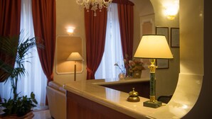Zona de recepción de un hotel con mostrador de mármol, lámpara dorada, cortina roja y candelabro.