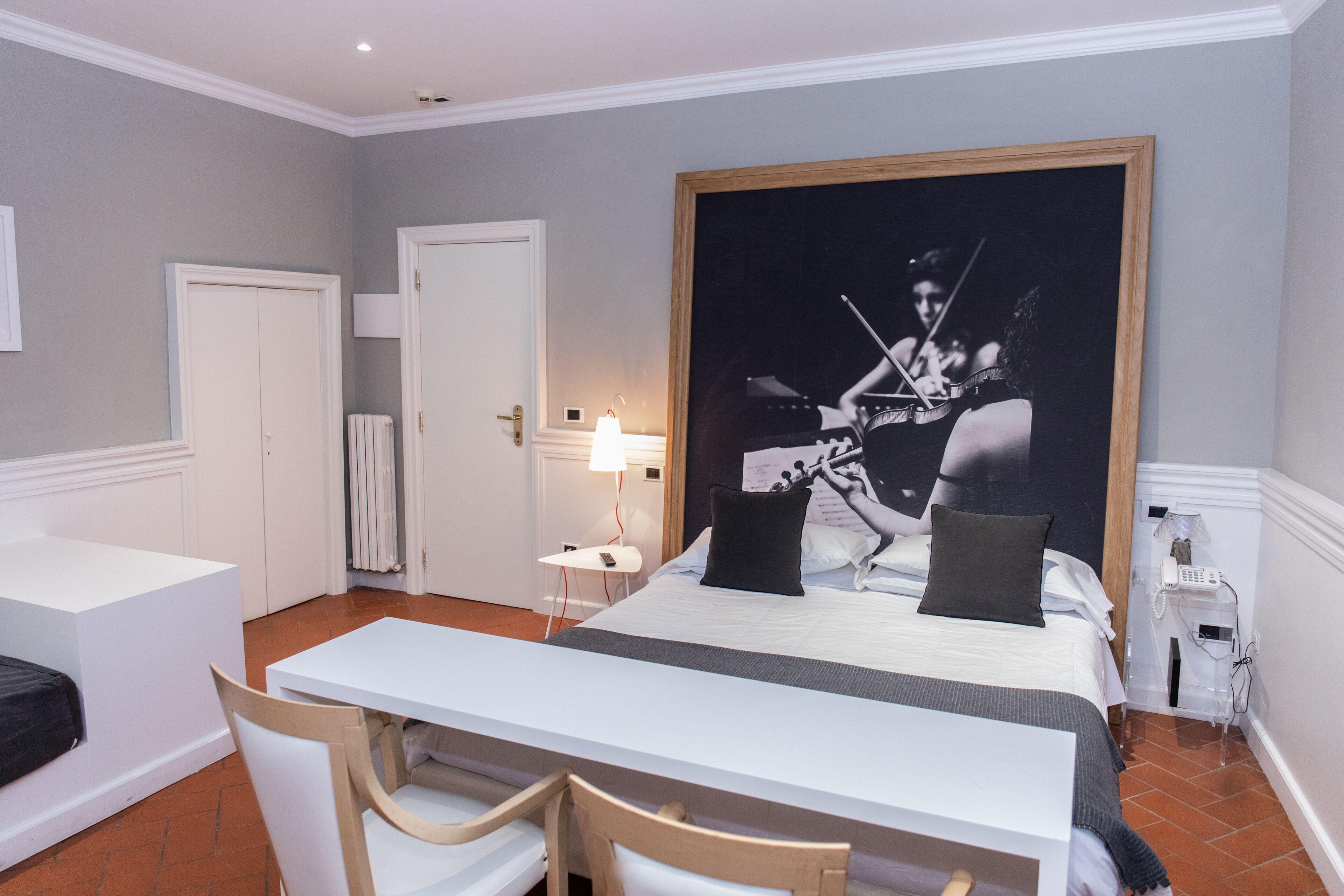 A hotel room with a bed, a desk, chairs, a large framed picture on the wall, a nightstand with a lamp, and a telephone.
