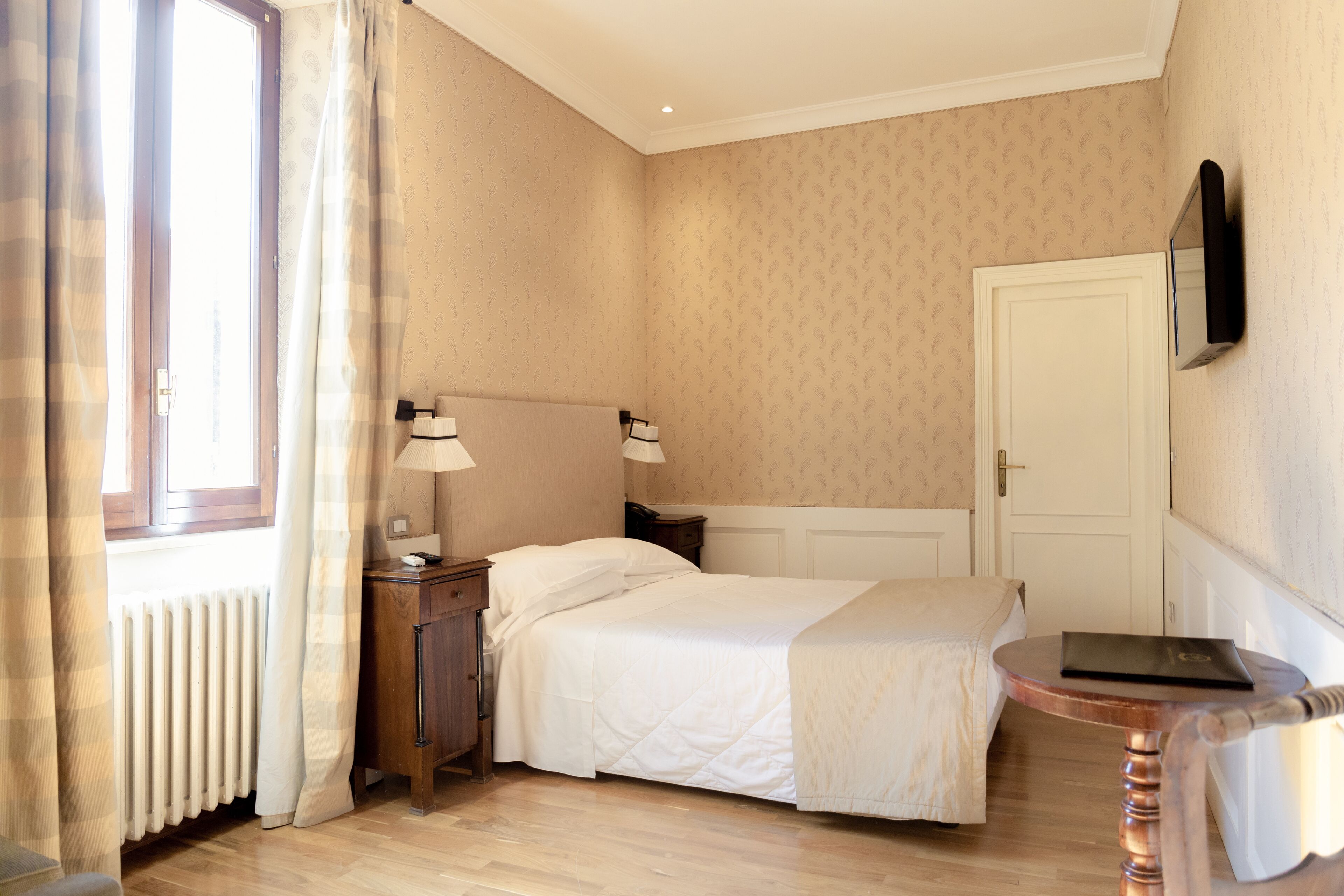 Quarto de hotel com cama, mesas de cabeceira, luminária, radiador, televisão na parede e janela com cortinas.