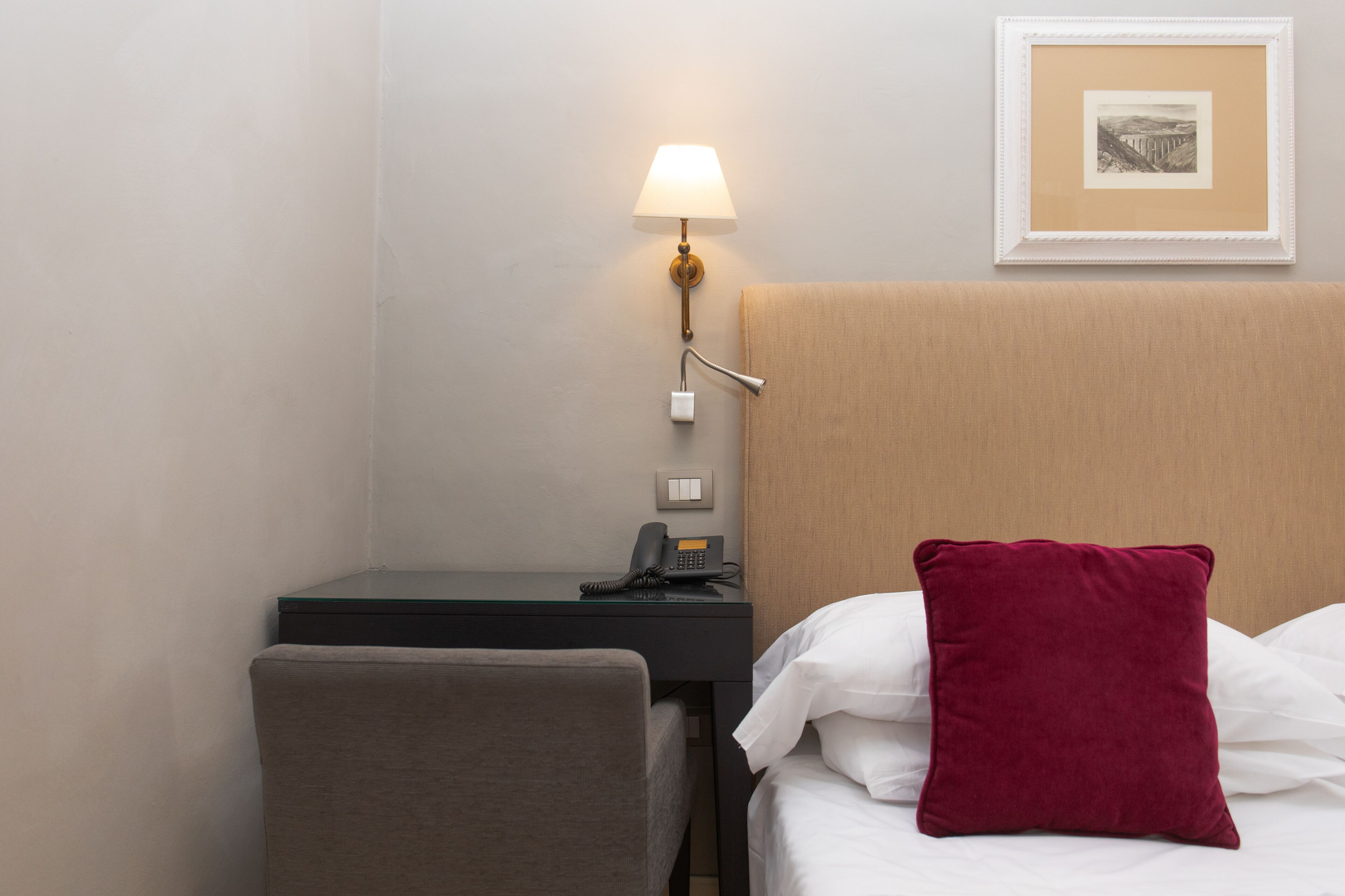 A hotel room with a bed, a desk, a chair, a lamp, a phone, and a framed picture on the wall.