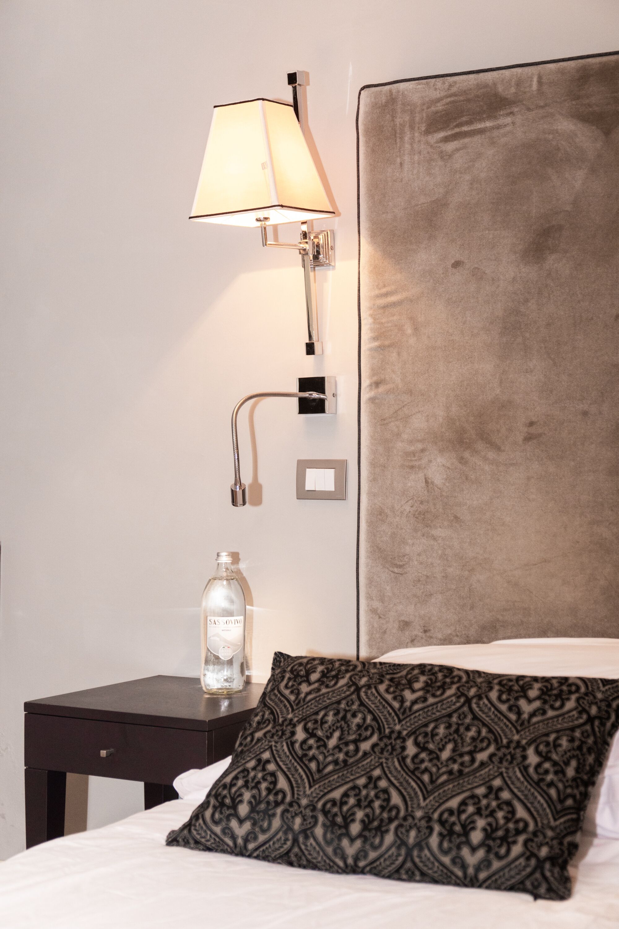 A neatly made bed with a patterned pillow, a bedside table with a bottle, and a wall-mounted lamp.