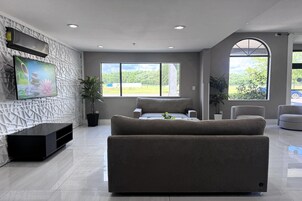 Una sala de estar moderna con un televisor de pantalla plana, un sofá, una silla y ventanas amplias con vistas al exterior.