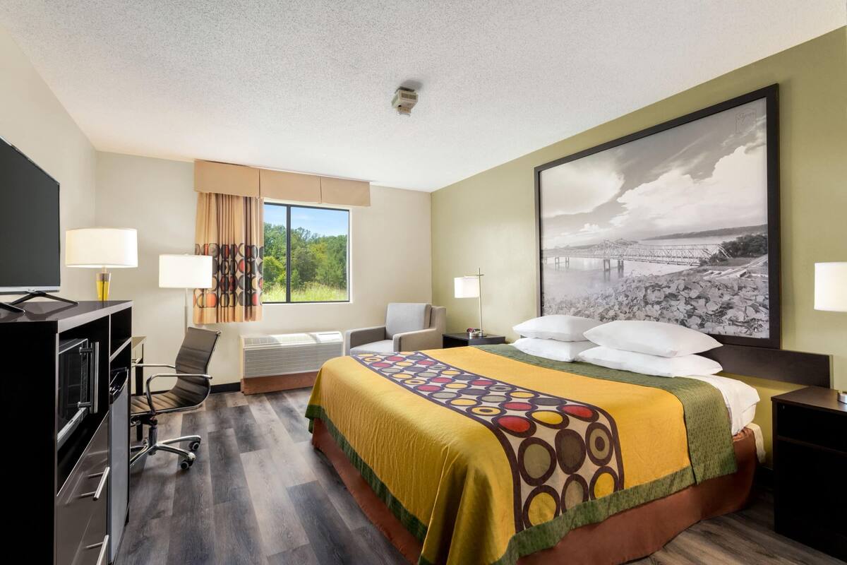 A hotel room with a bed, a desk, a chair, a TV, and a window with a view of greenery.