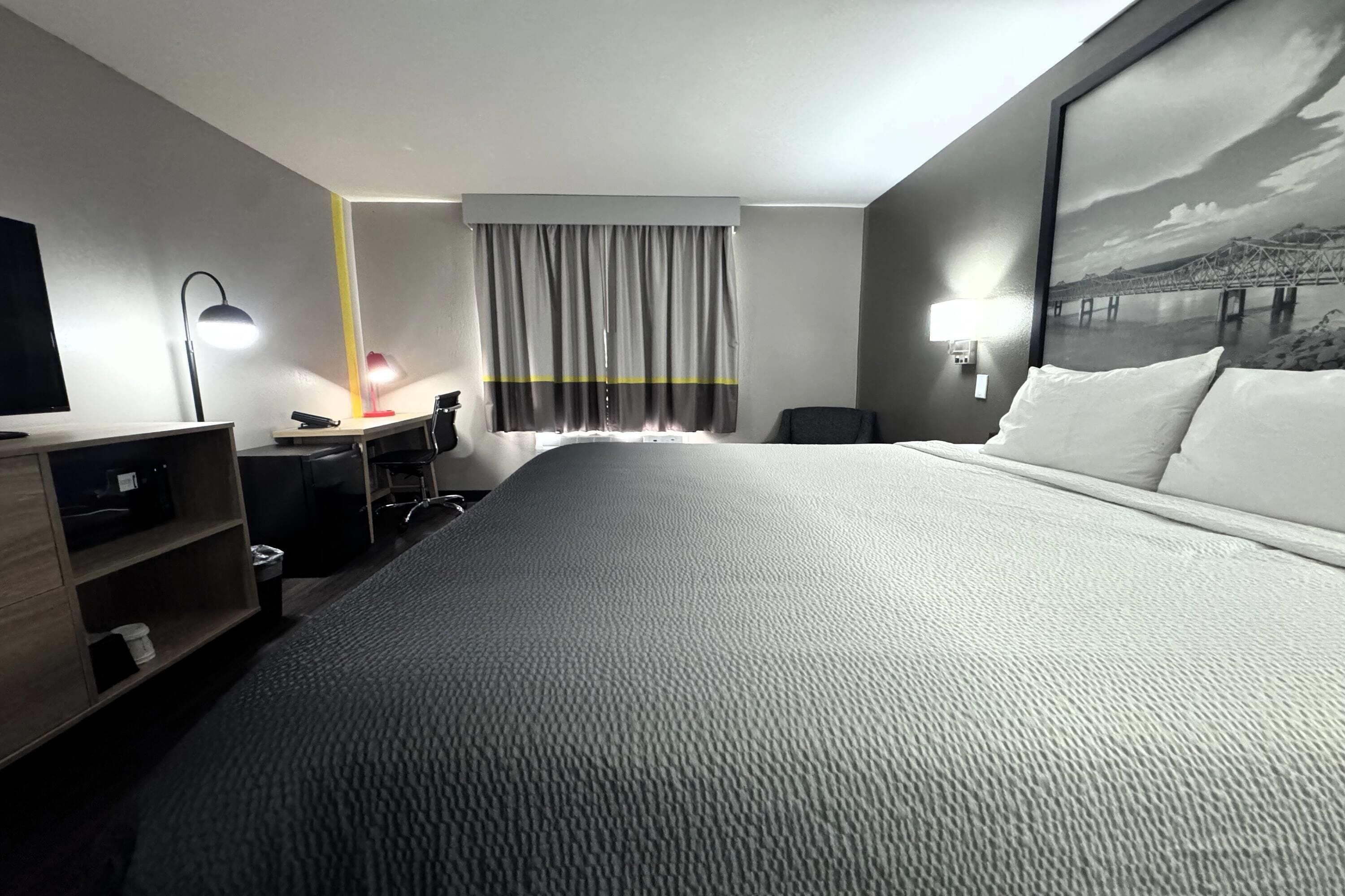 Una camera d'albergo con un letto grande, una scrivania con una sedia, una lampada da scrivania, un cestino e una finestra con tende.
