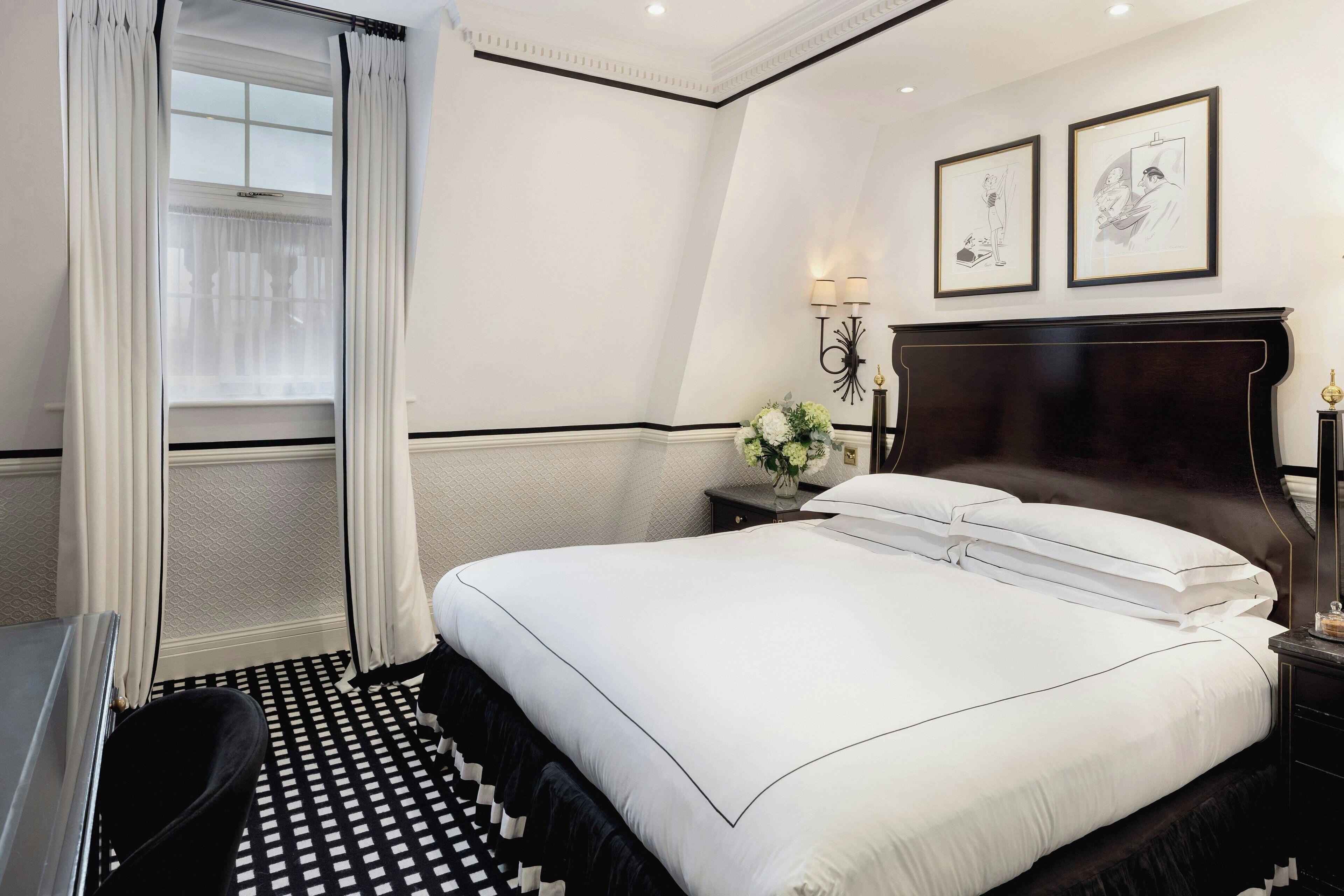 A hotel room with a large bed, a desk, and a chair. There are two framed pictures on the wall, a nightstand with a lamp, and a window with curtains.