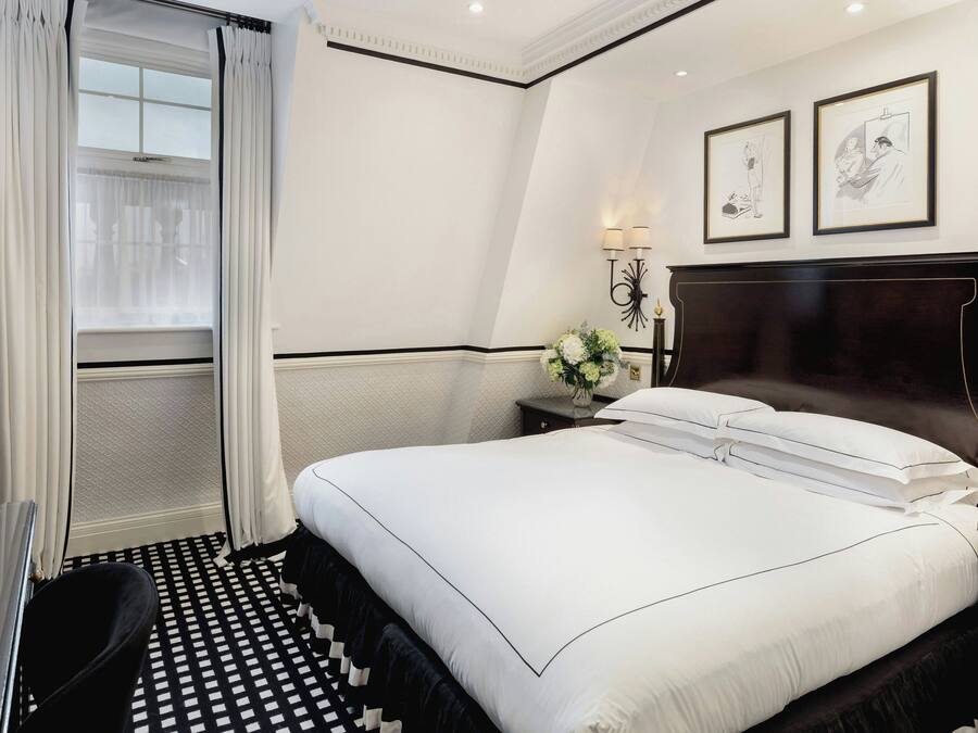 A hotel room with a large bed, a desk, and a chair. There are two framed pictures on the wall, a nightstand with a lamp, and a window with curtains.