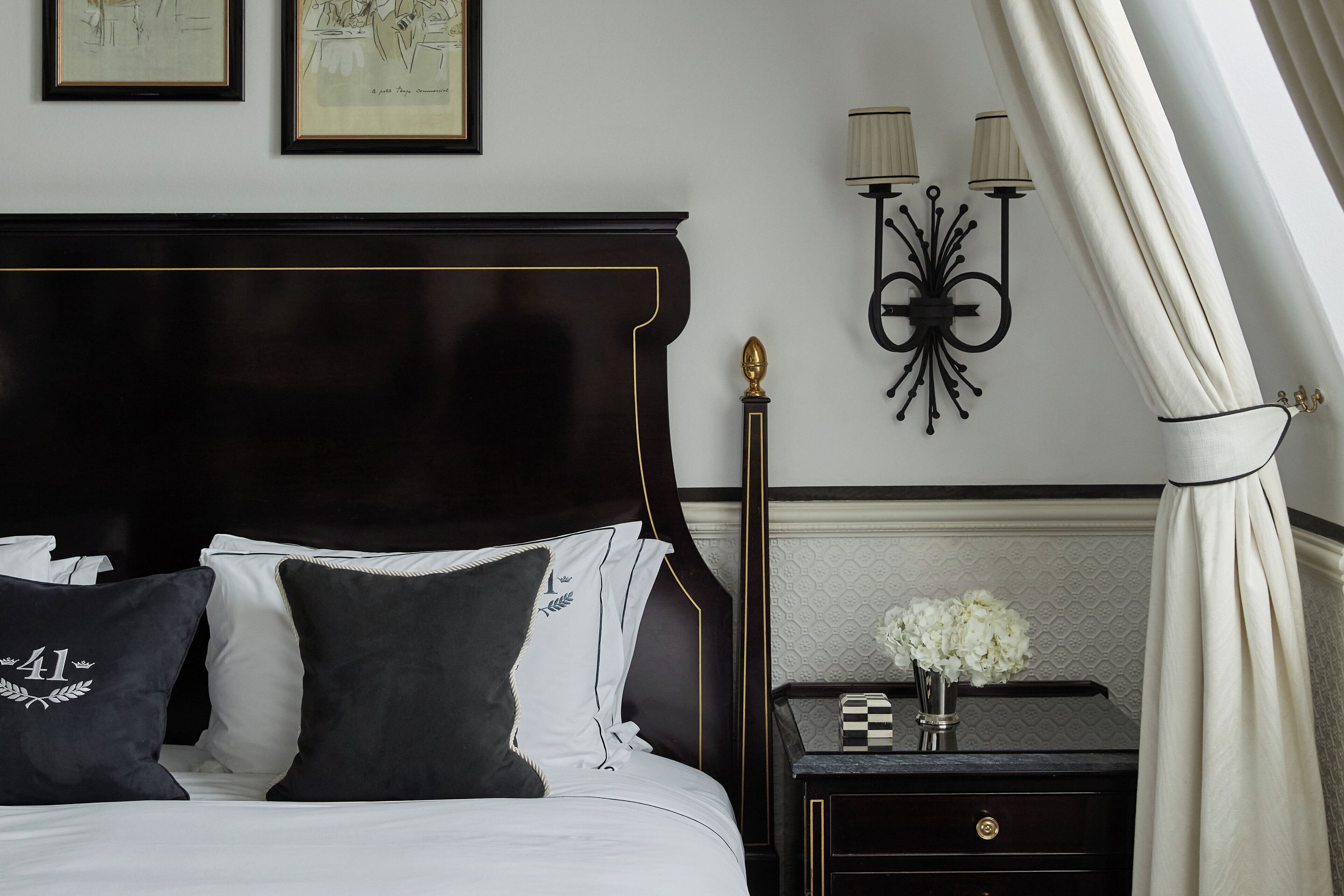 Una cama bien tendida con ropa de cama blanco y negro, una mesita de noche con un jarrón de flores y una lámpara de pared.