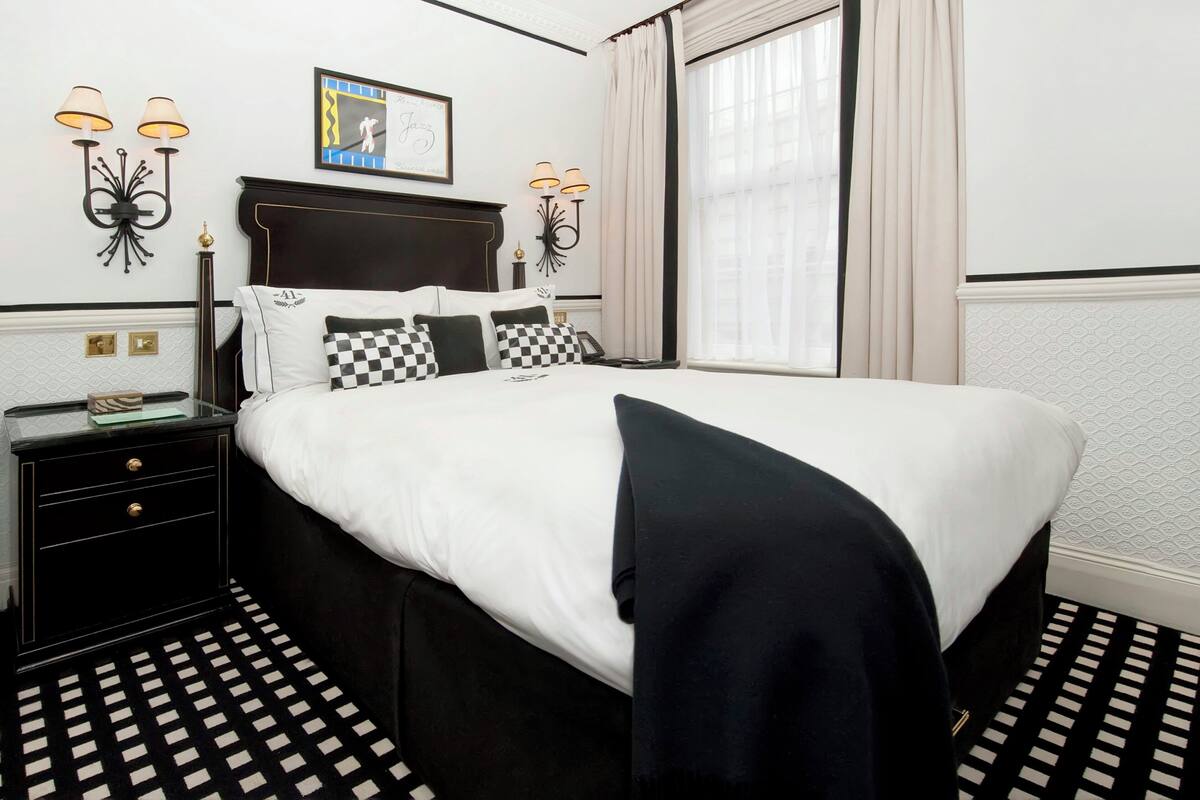 A hotel room with a large bed, two bedside tables, a nightstand, a wall-mounted lamp, and a window with curtains.