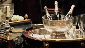 Ein Mann im Anzug gießt Champagner in ein Kristallglas. Auf einem Tablett befinden sich Eiswürfel, zwei Champagnerflaschen und mehrere Gläser.