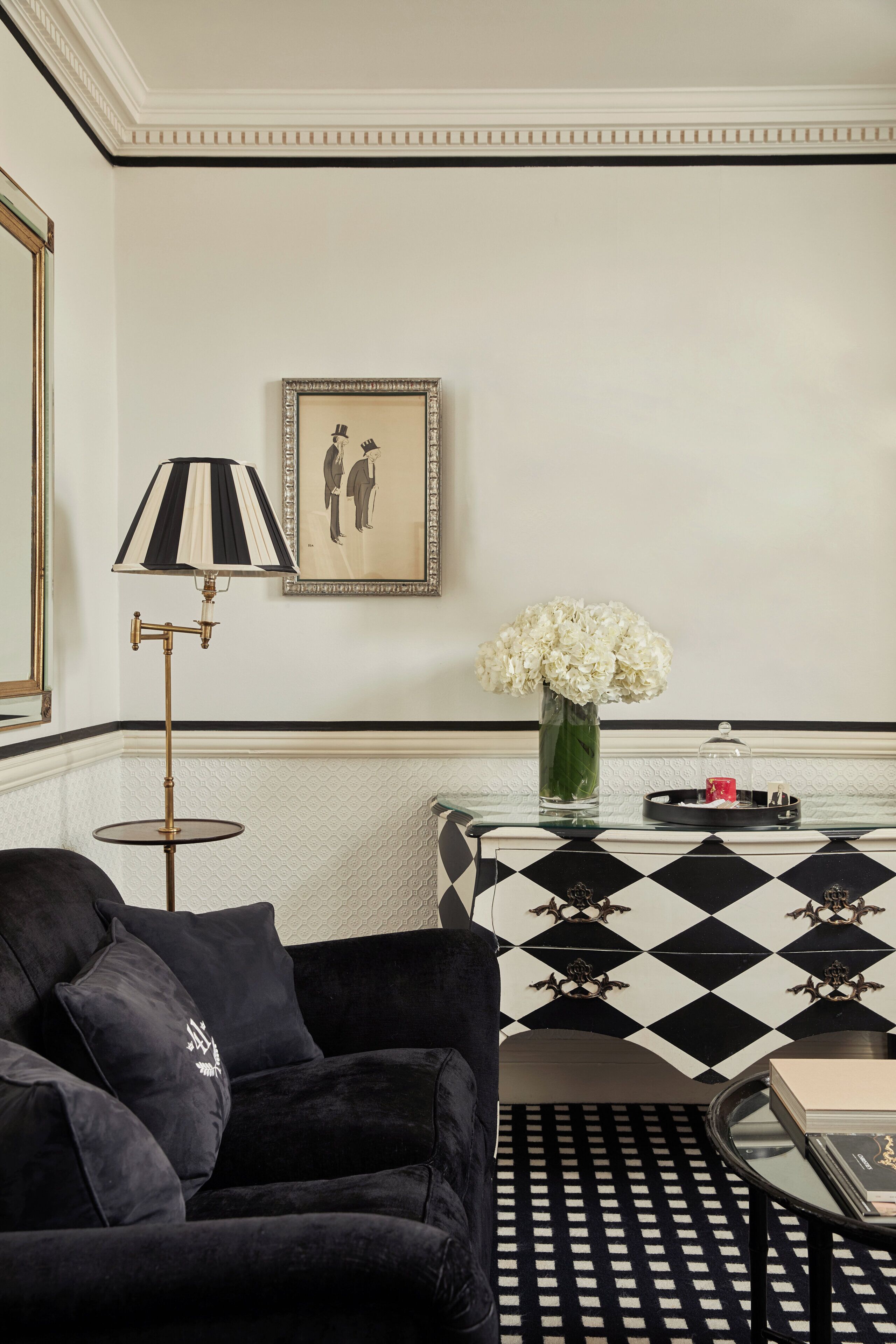 Un canapé en velours noir, une table à motifs, une lampe sur pied, un vase avec des fleurs blanches et un tableau encadré au mur.