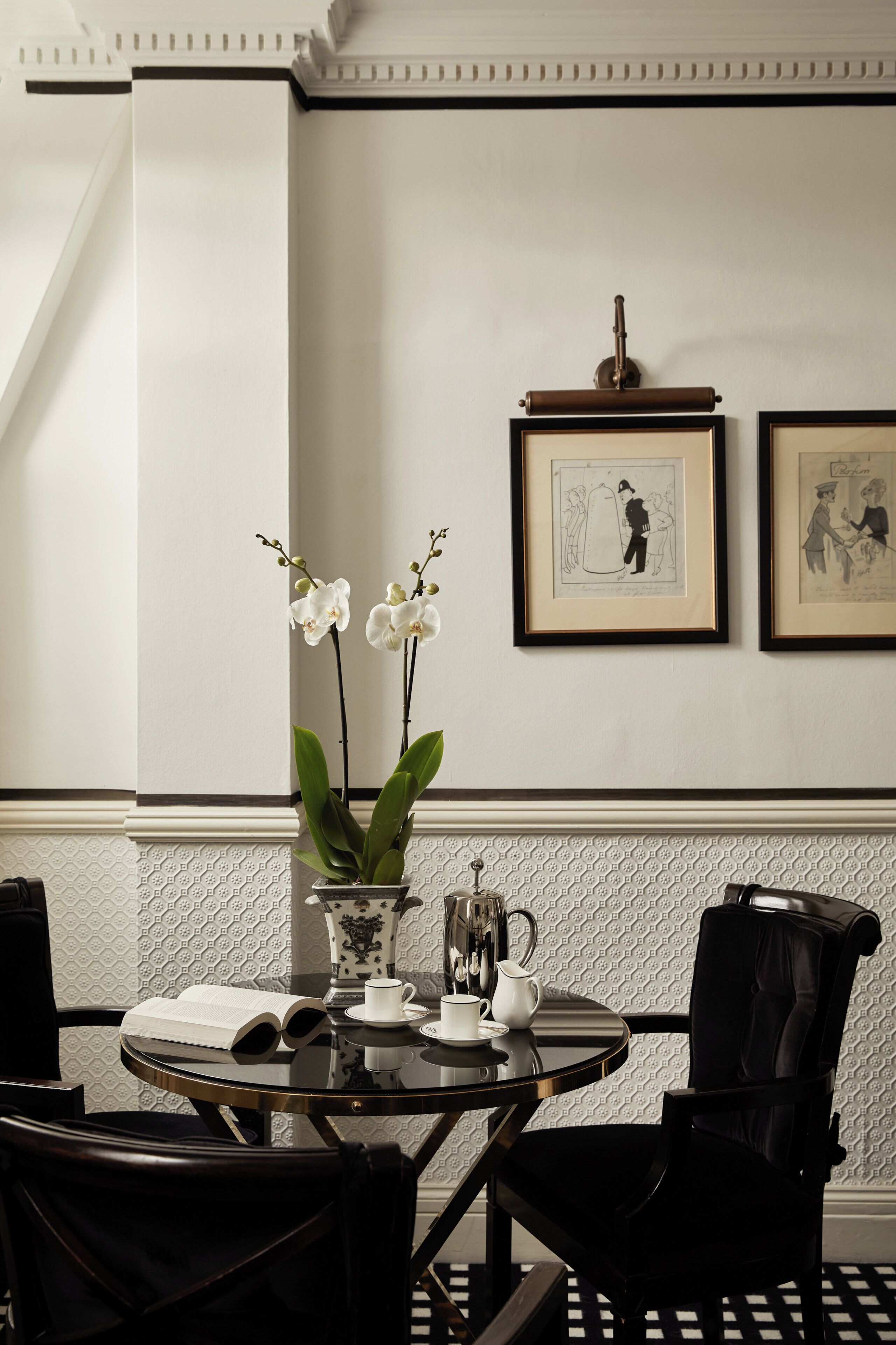 Un coin repas avec une table ronde, des chaises noires, un vase contenant des orchidées blanches, des tableaux encadrés au mur et un papier peint à motifs.