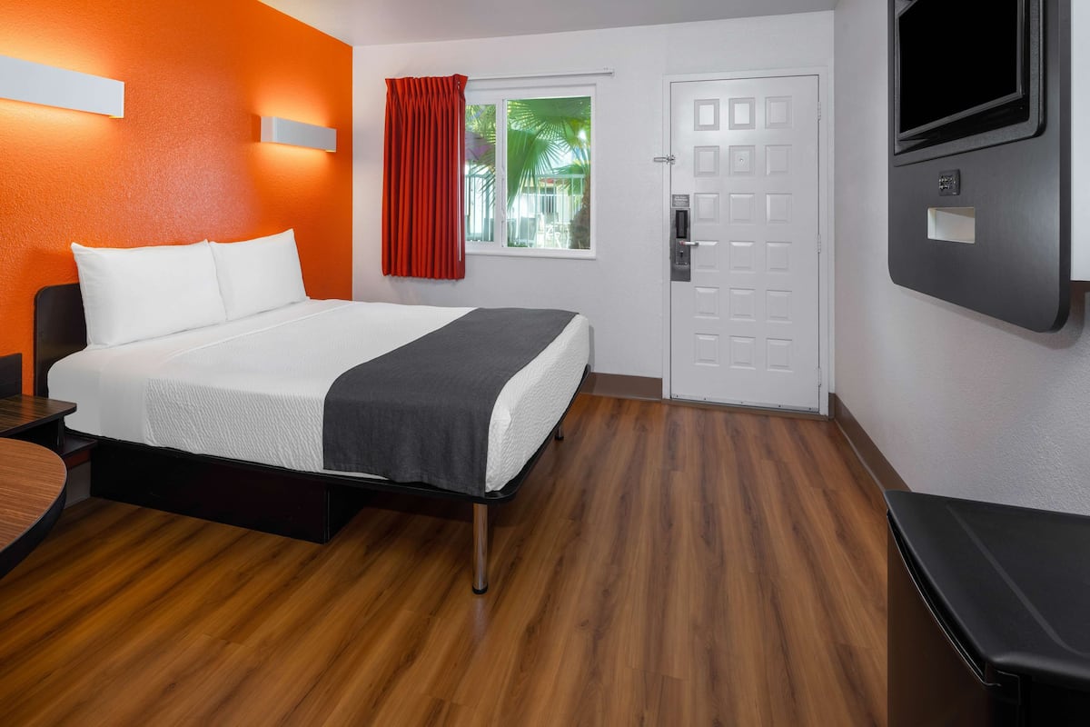 A hotel room with a bed, a desk, a TV, a window with curtains, and a door.