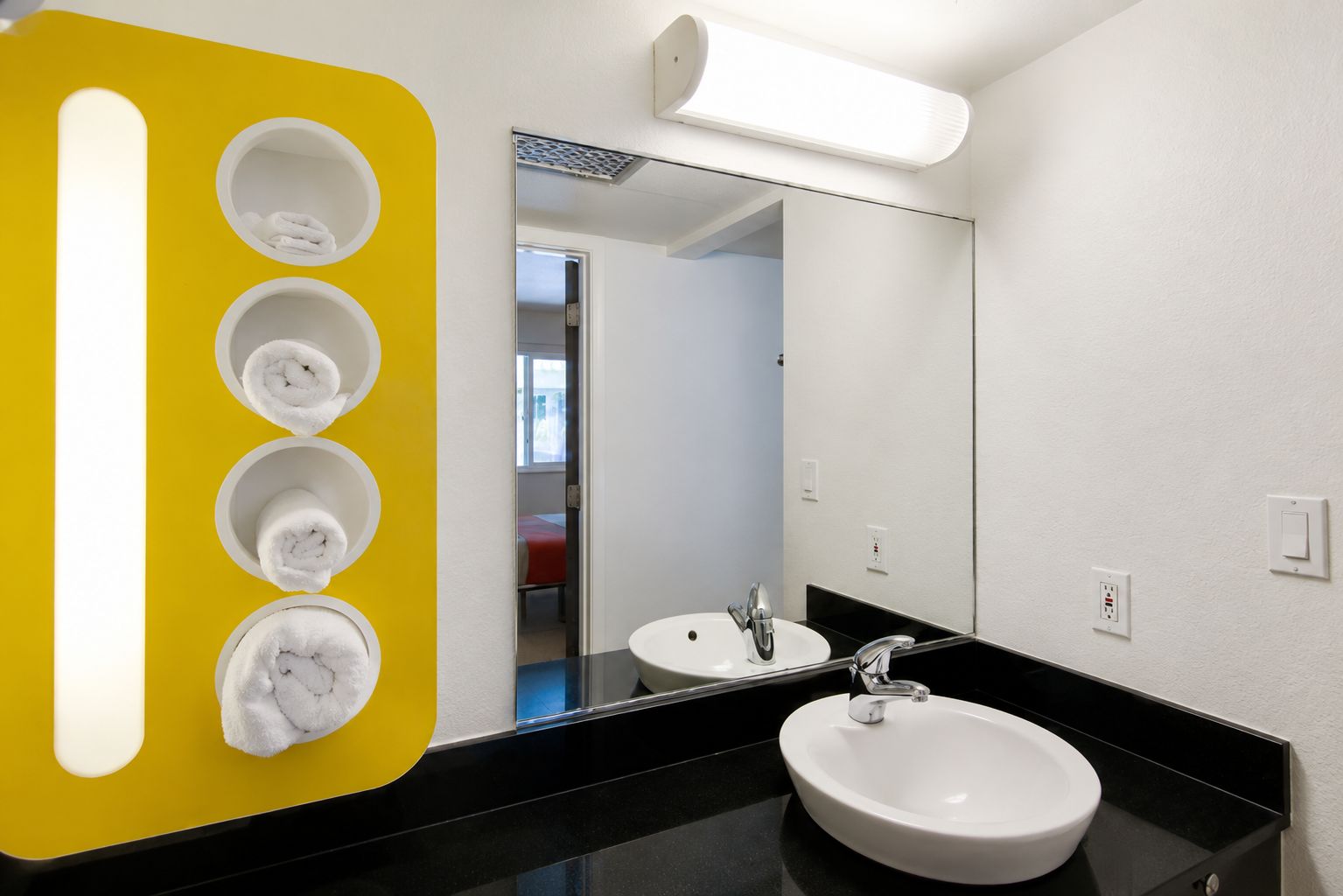 Un baño moderno con un mostrador negro, un lavamanos blanco y un espejo. Hay cuatro toallas blancas colgadas en una pared amarilla.
