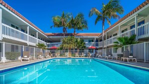 A swimming pool with lounge chairs and palm trees, surrounded by a building with balconies and red awnings.
