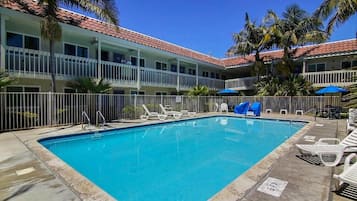 A swimming pool with lounge chairs and umbrellas, surrounded by a building with balconies and palm trees.
