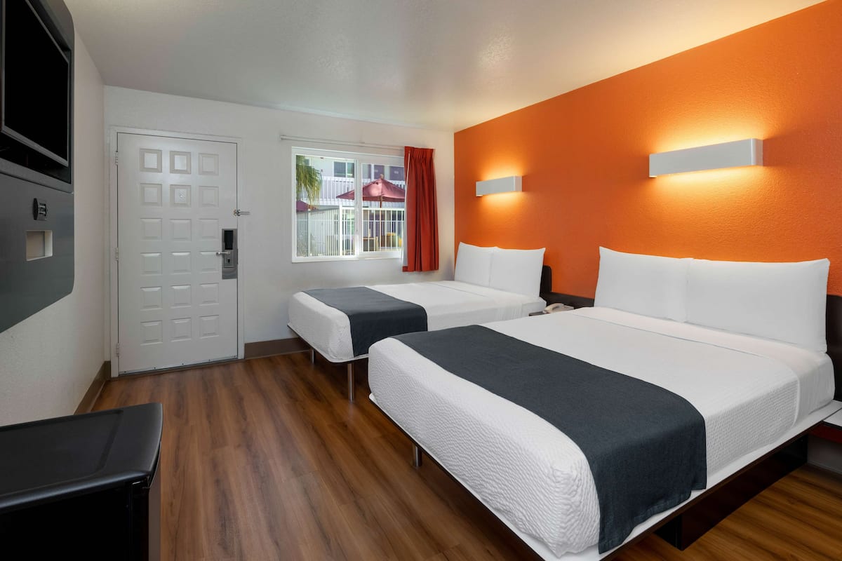 A hotel room with two beds, a TV, a window with curtains, and an orange accent wall.
