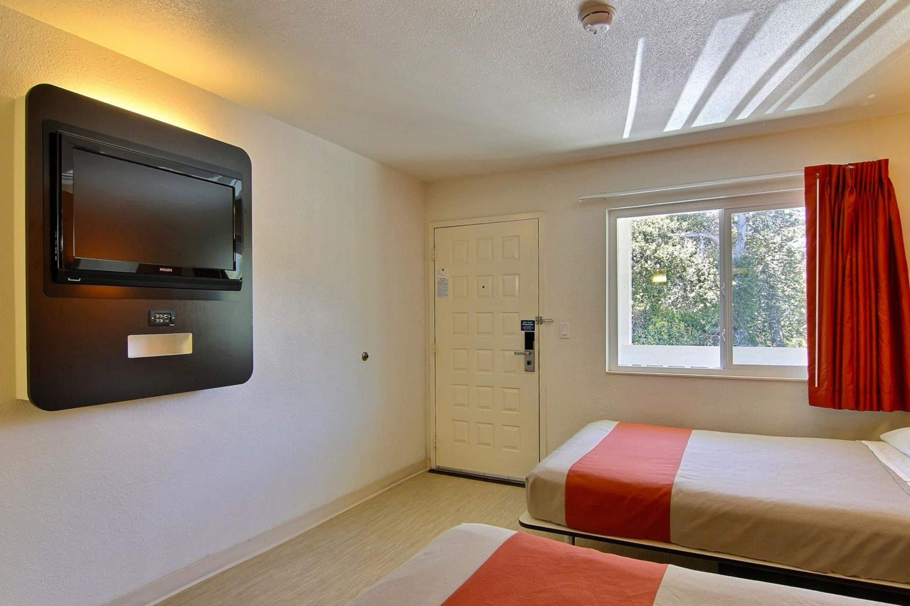 A hotel room with a bed, a television mounted on the wall, a window with curtains, and a door.