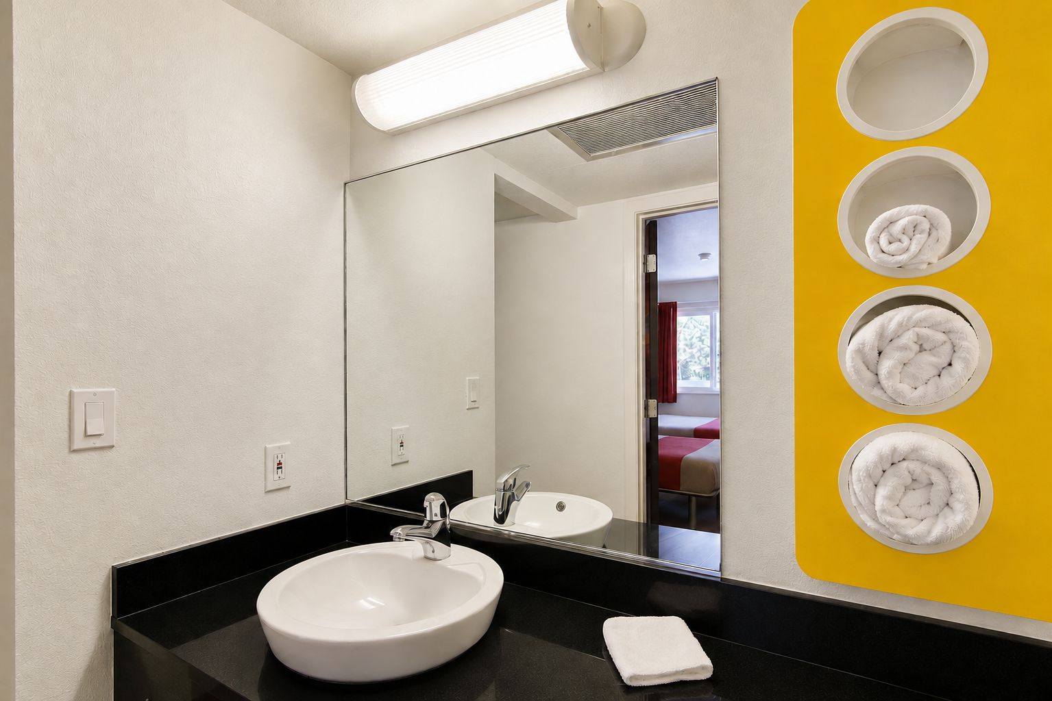 A modern bathroom with a large mirror, a white sink, and a black countertop. There are four towel hooks on the wall.