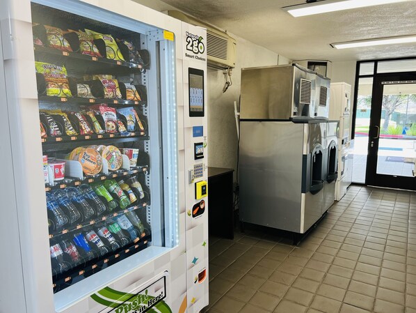 Máquina expendedora con aperitivos y bebidas, una cafetera y un refrigerador en un entorno comercial.