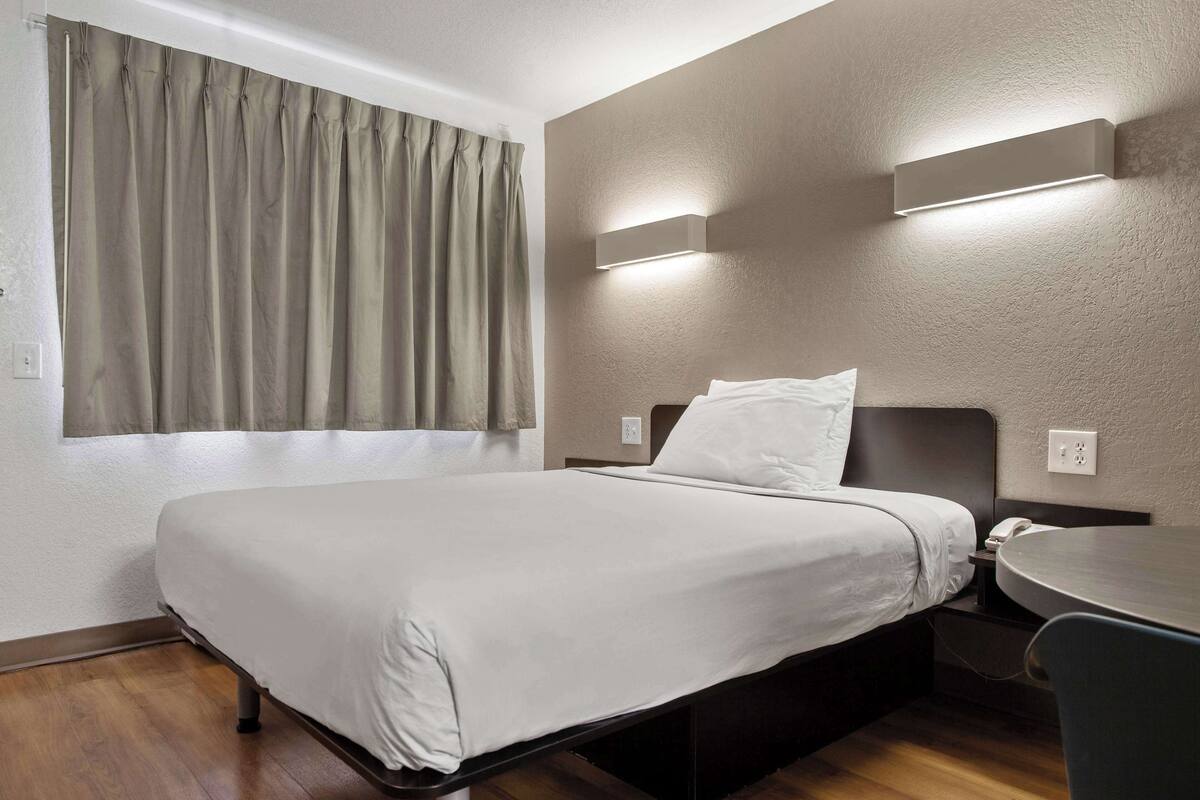 A hotel room with a bed, a bedside table, a chair, and a window with curtains.