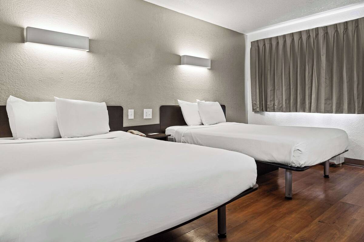 A hotel room with two beds, a bedside table with a lamp, a wall-mounted light, and a window with curtains.