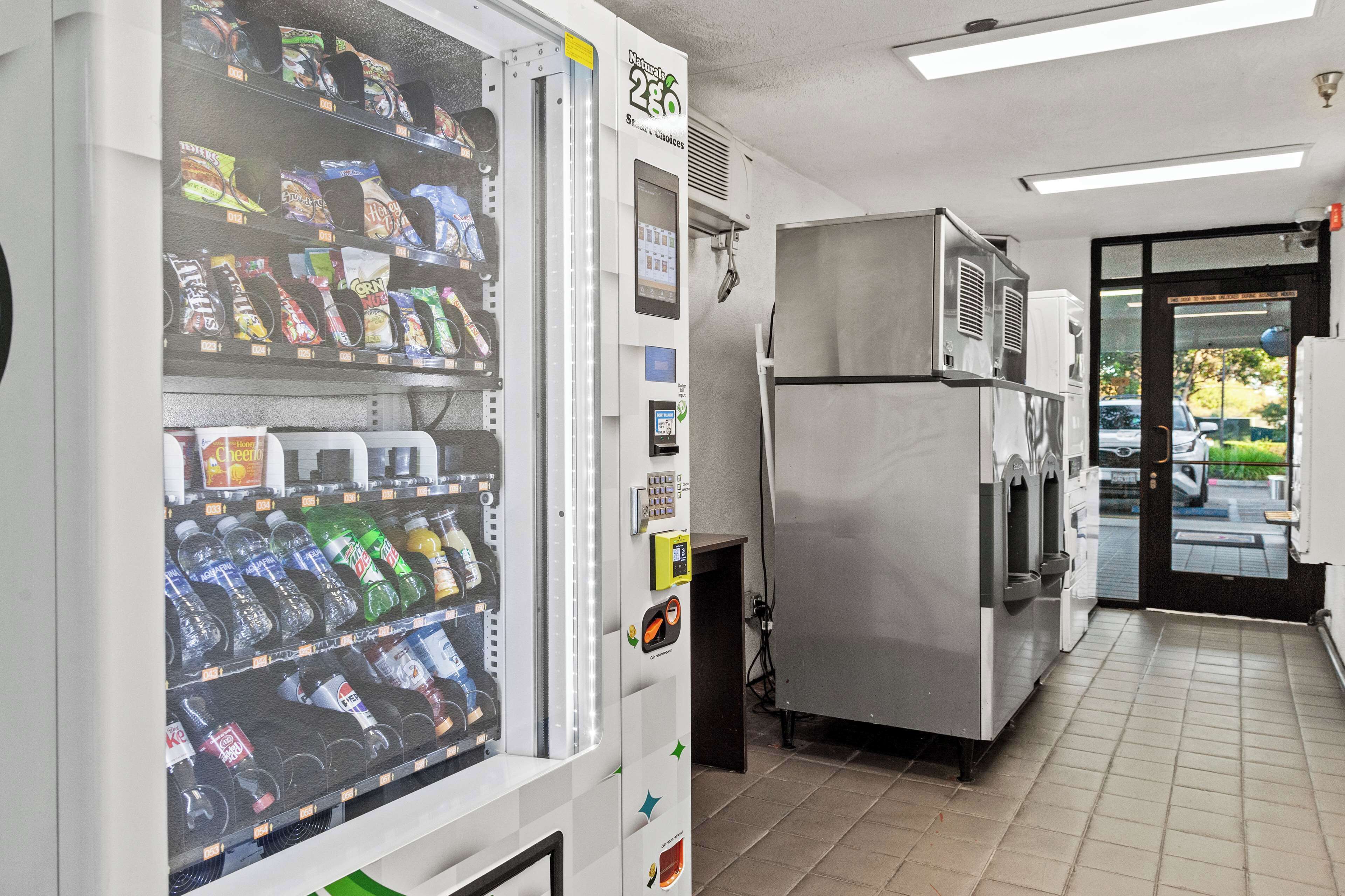 A vending machine stocked with snacks and drinks, a stainless steel refrigerator, and a tiled floor in a commercial setting.