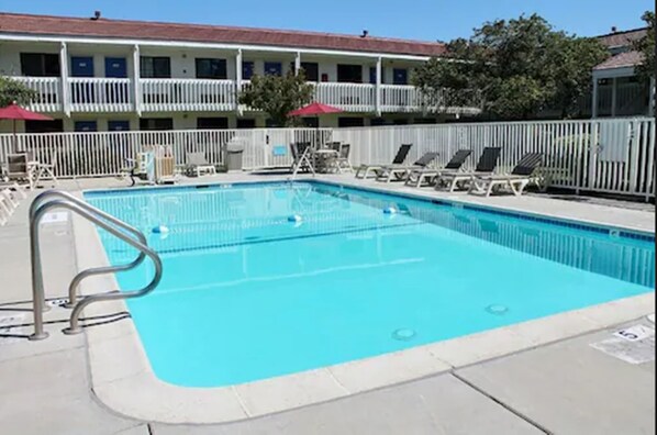 Una piscina rectangular con una escalera metálica en un lateral, rodeada por una valla blanca. Hay sillas de descanso a lo largo de la valla y un edificio con balcones al fondo.