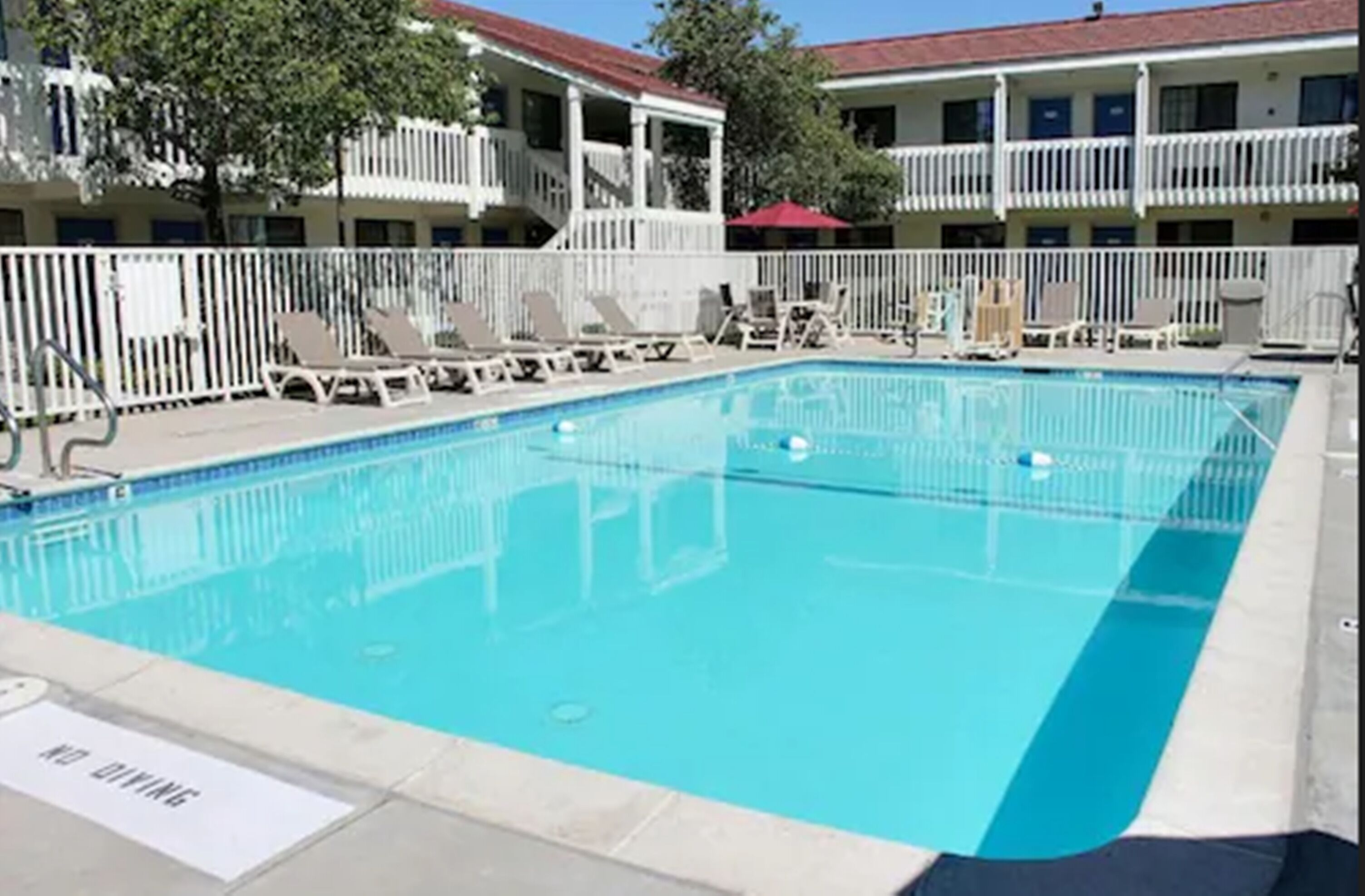 A rectangular swimming pool with lounge chairs and umbrellas, surrounded by a white fence and a building with balconies.