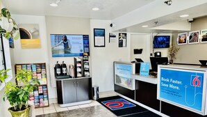 A hotel reception area with a digital screen displaying a booking message, a coffee machine, a plant, and informational posters on the walls.