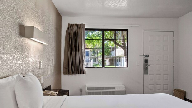 A hotel room with a bed, a window with curtains, a wall-mounted light, and a door.