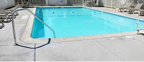 A rectangular swimming pool with a white deck and a metal ladder. There are lounge chairs and umbrellas around the pool.