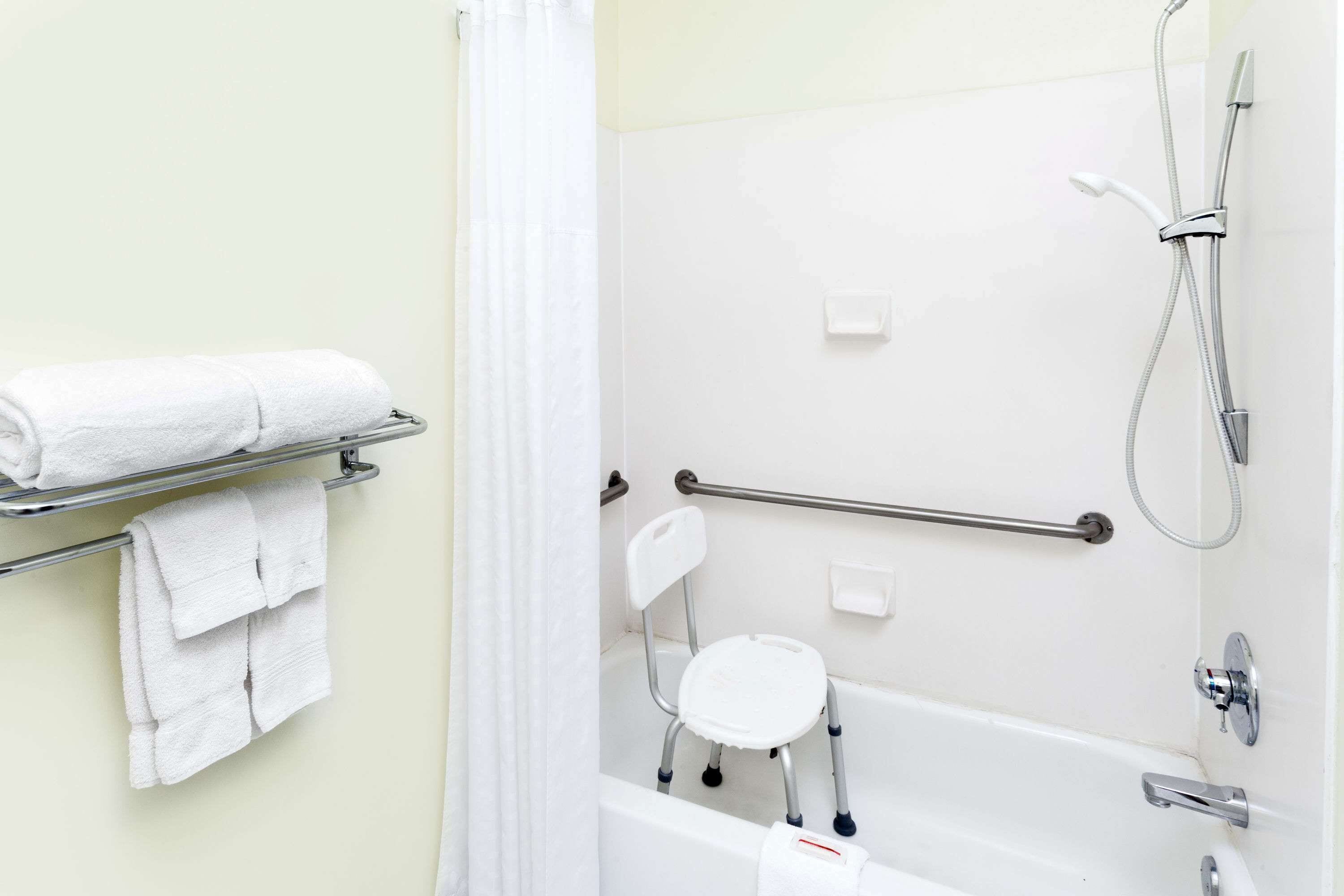 Een badkamer met een wit bad, een douche, een handgreep, een witte stoel en een handdoekrek met handdoeken.
