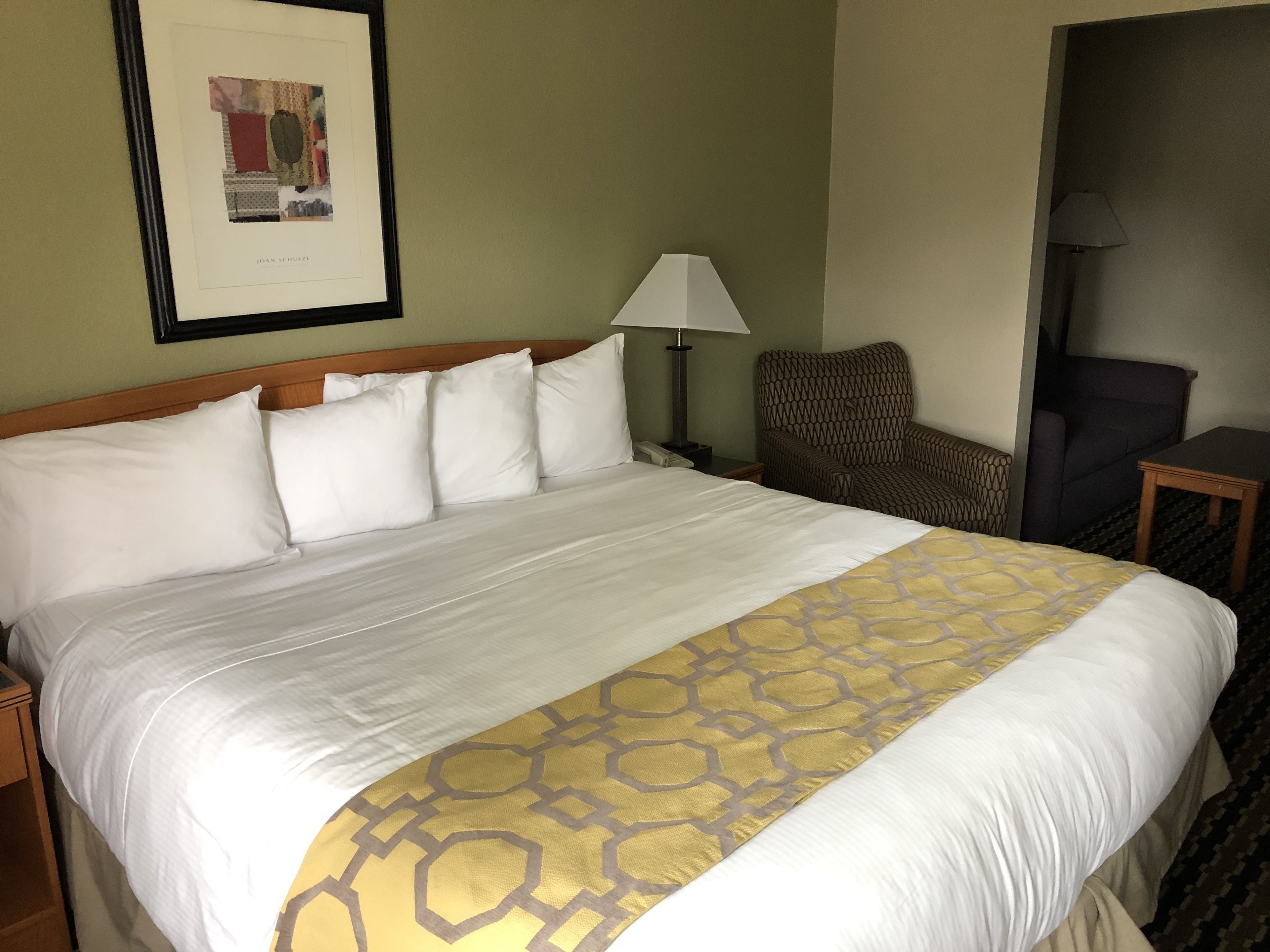 A neatly made bed with white and gold bedding, a framed picture on the wall, a lamp, and a chair.