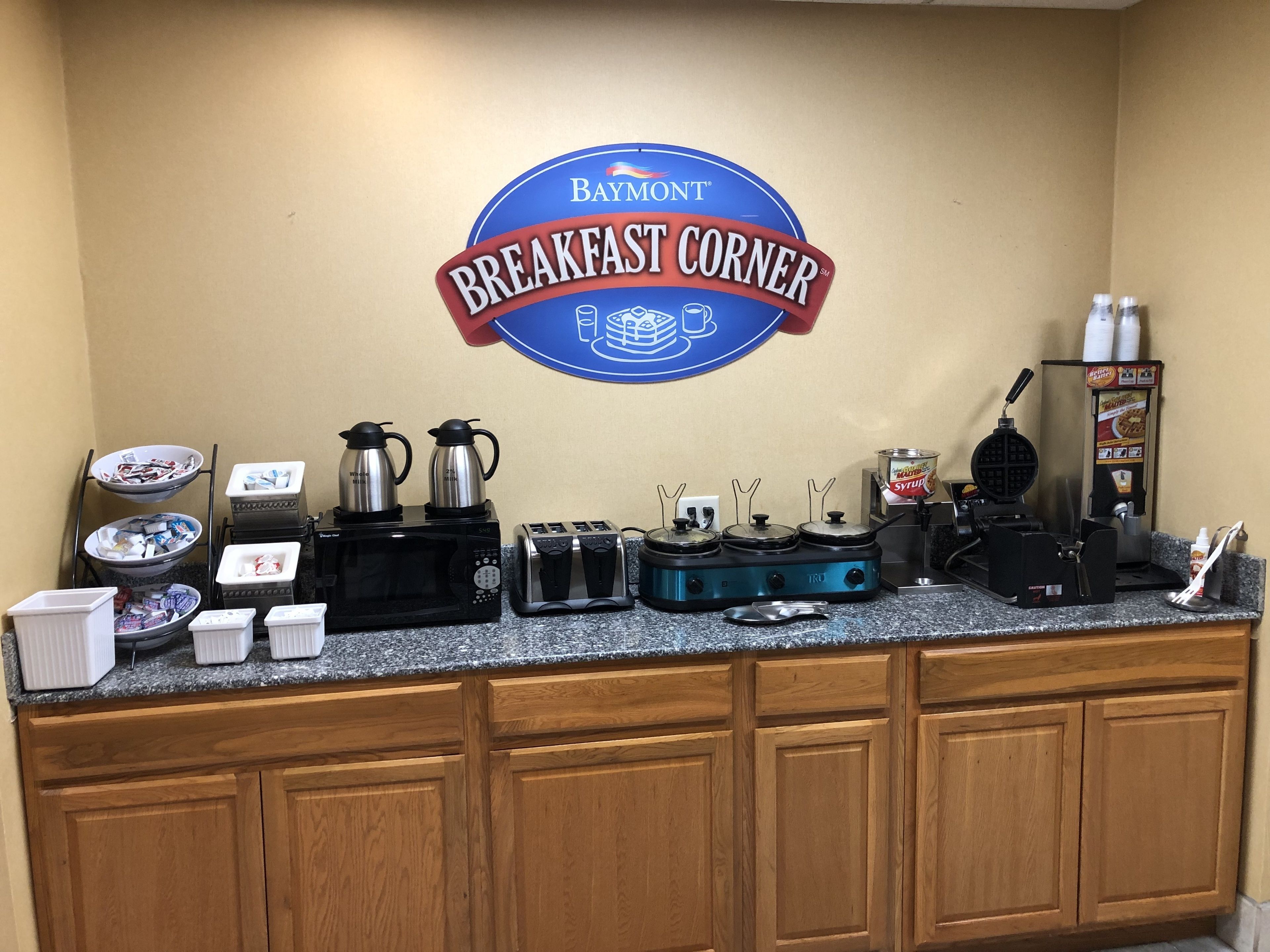 Ein Frühstücksbuffet mit einem Schild ‚Baymont Breakfast Corner‘, einer Kaffeemaschine, einem Waffelautomat und einem Sirup-Spender.