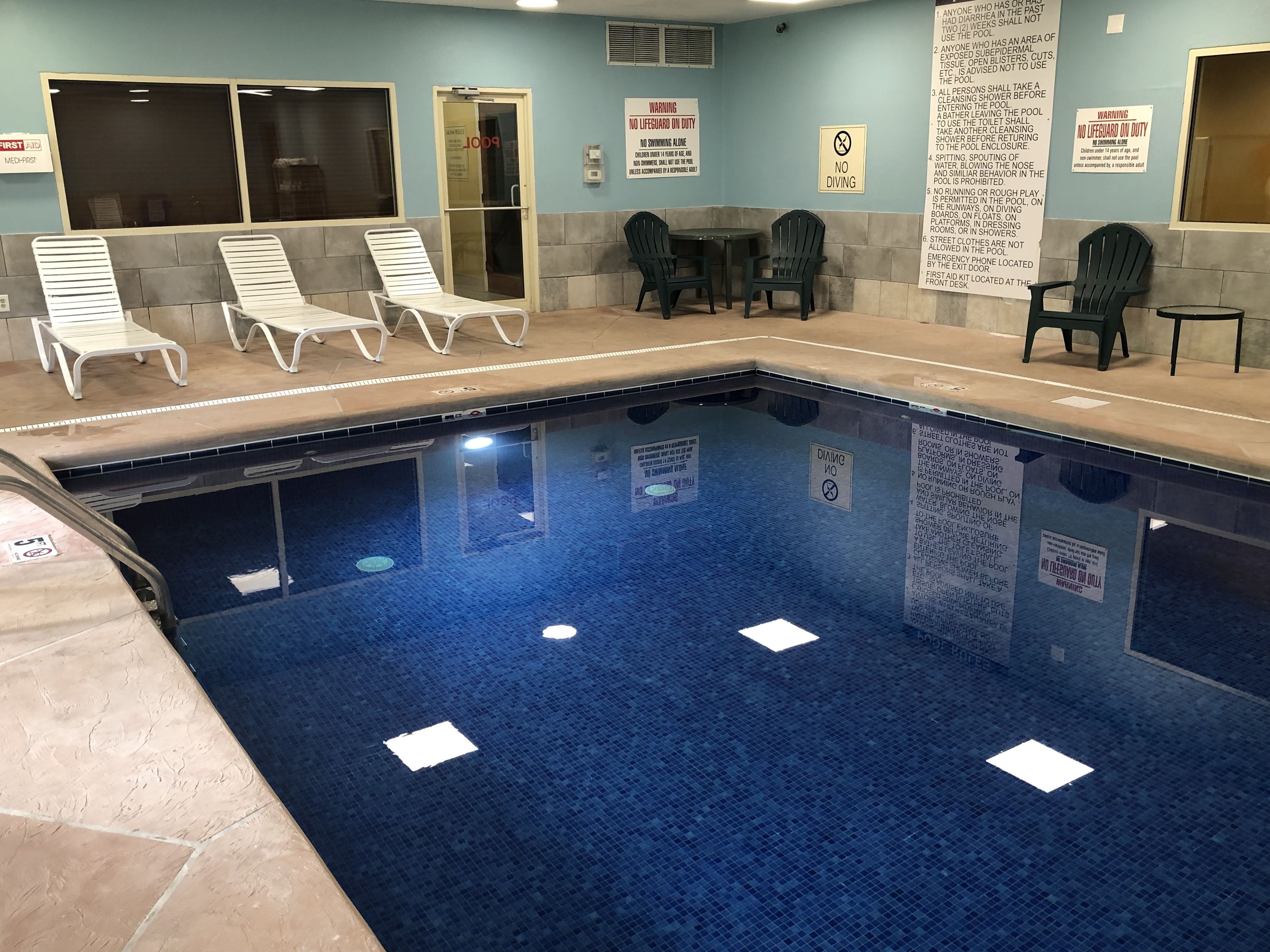 Une piscine intérieure équipée de chaises longues, d’une table et de divers panneaux d’information relatifs à la sécurité et aux règles d’utilisation.