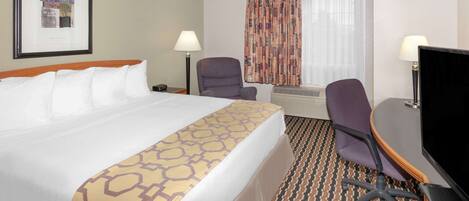 A hotel room with a large bed, a desk with a computer, a chair, a TV, and a window with curtains.