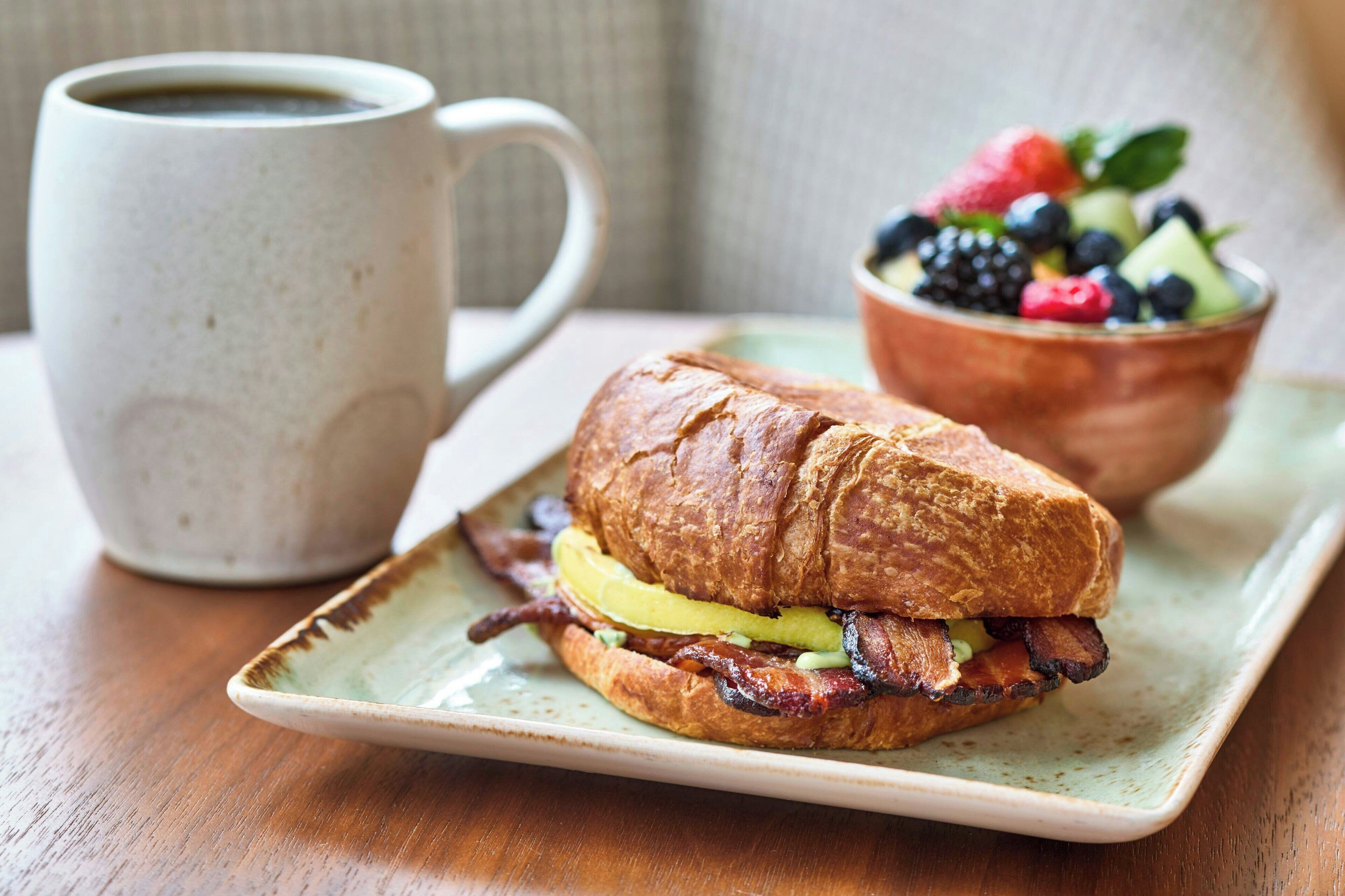 Un sándwich con tocino, una taza de café y un bol de fruta en un plato.