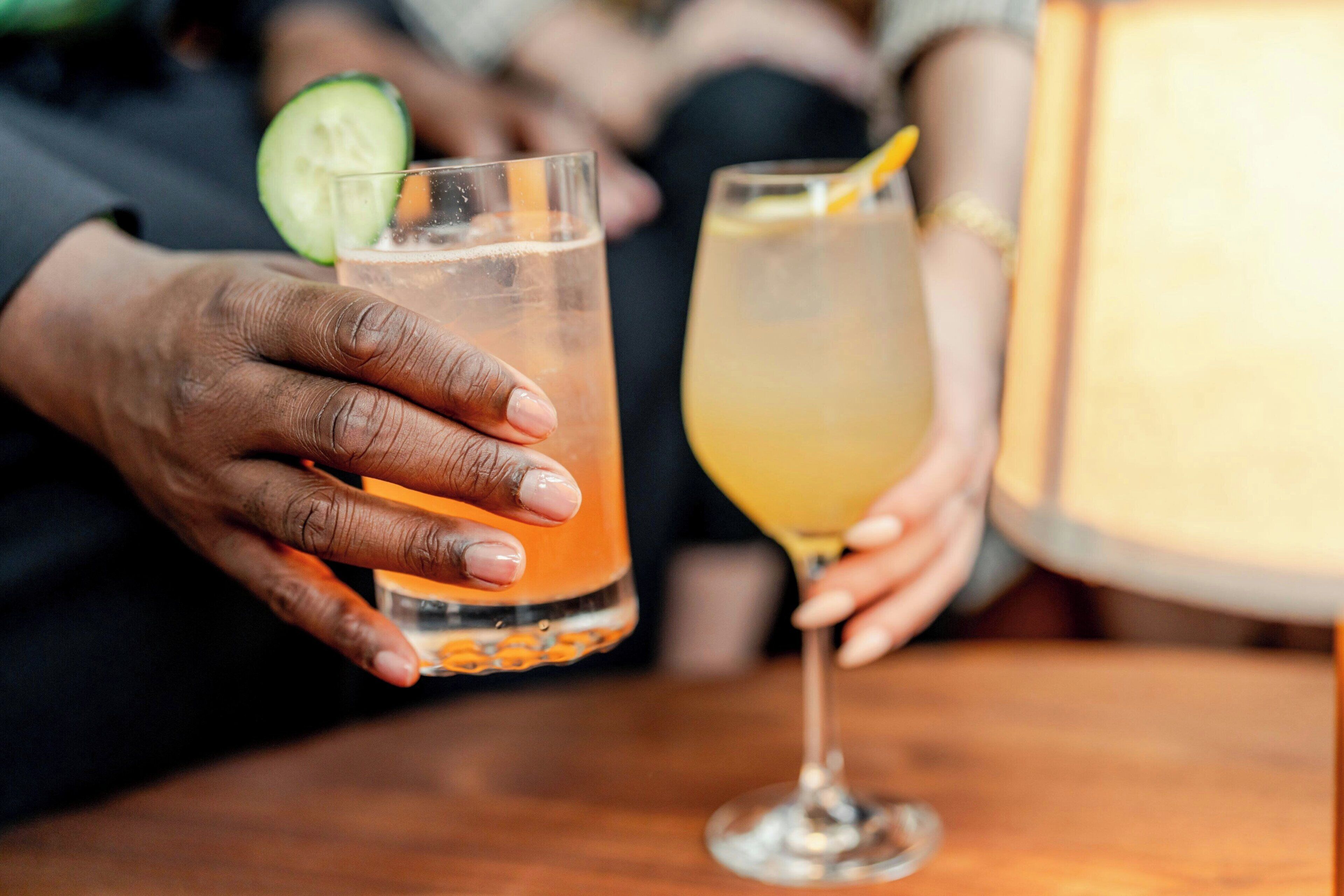 Un gros plan de deux mains tenant deux verres remplis de boissons, l’un contenant une tranche de concombre et l’autre une tranche de citron.
