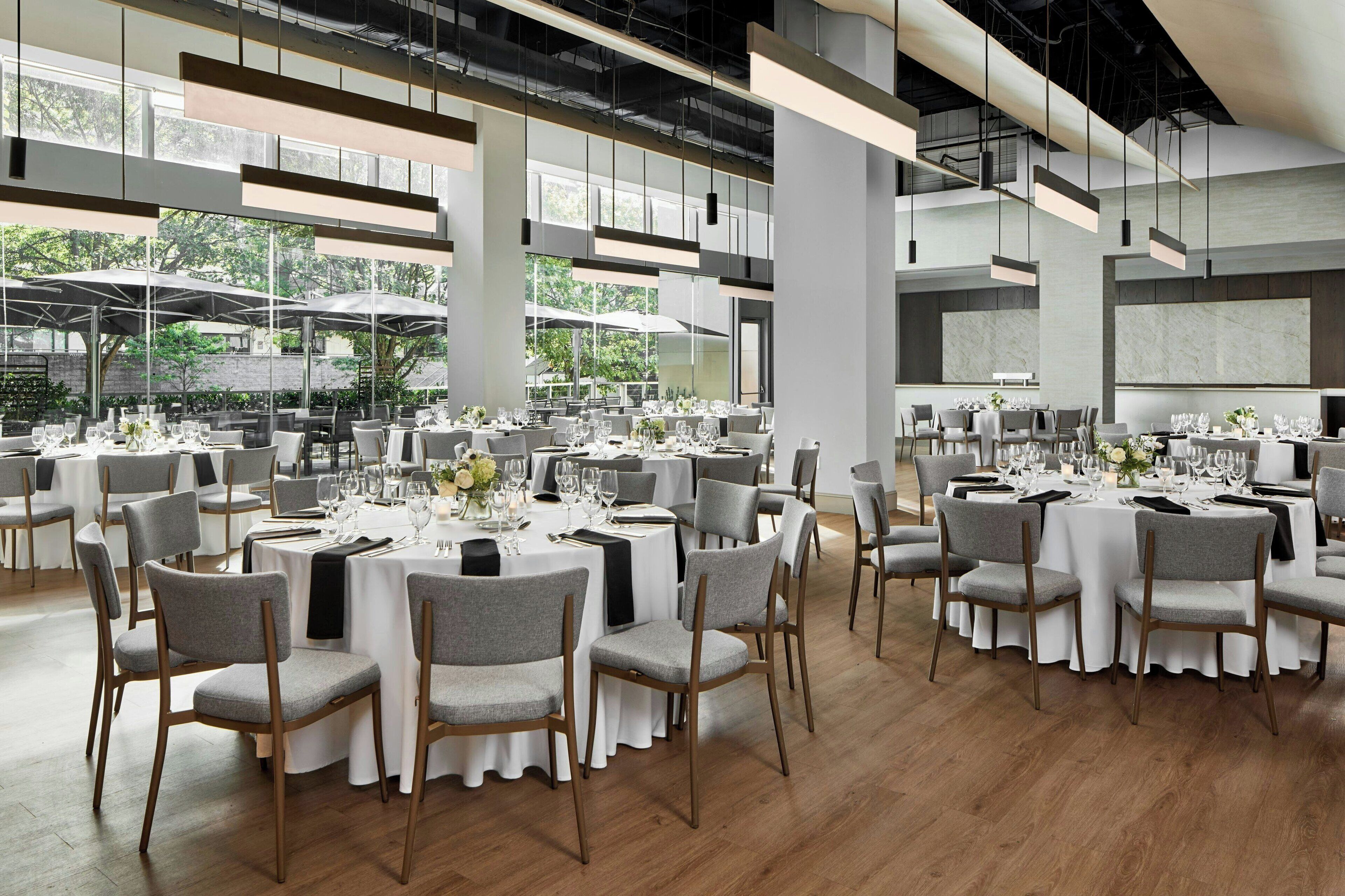 Un comedor amplio con mesas redondas y sillas tapizadas grises, ventanas grandes que ofrecen vistas al exterior y un moderno diseño de techo industrial.
