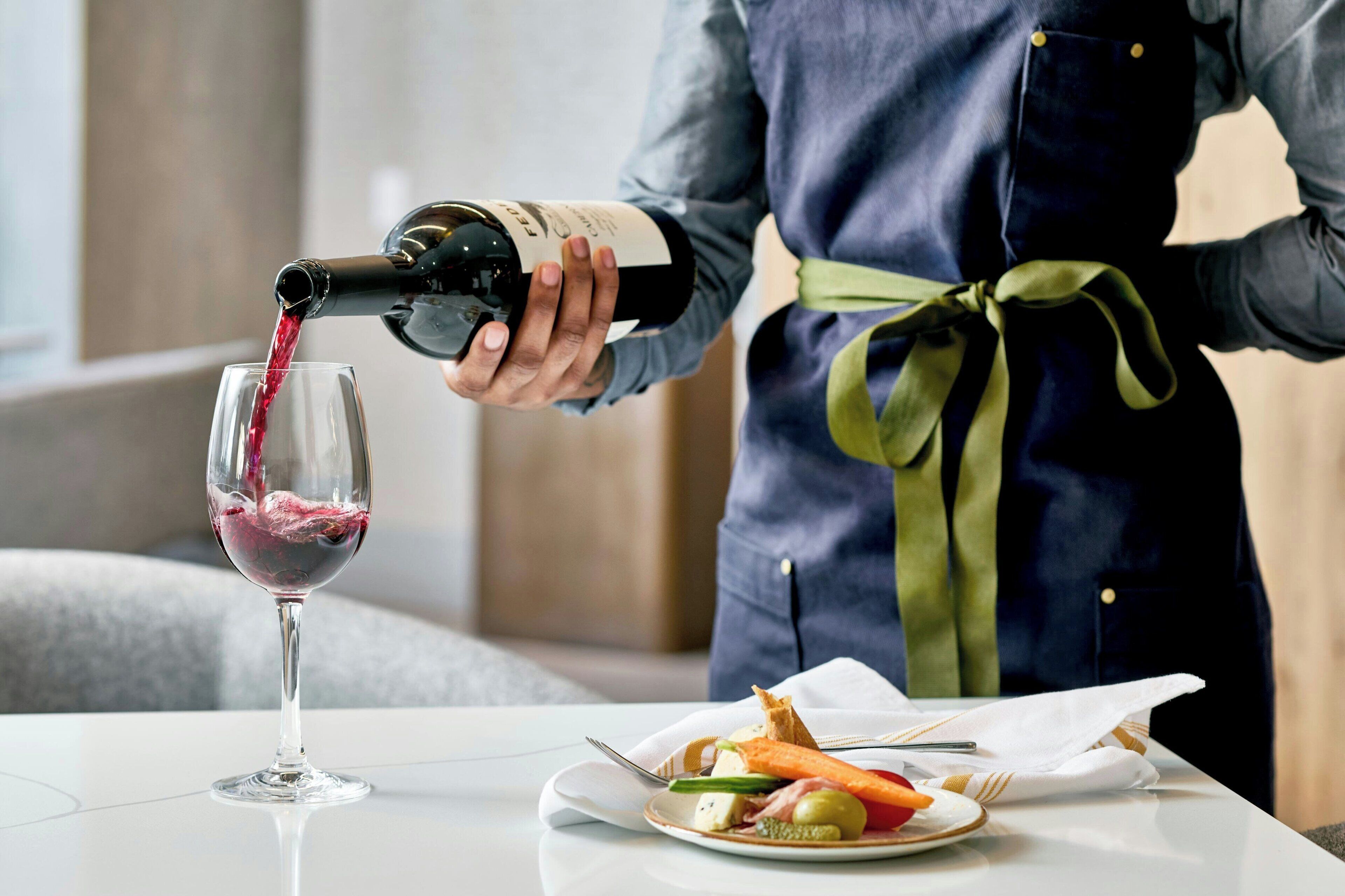 A person pouring red wine into a glass, standing next to a plate with food.