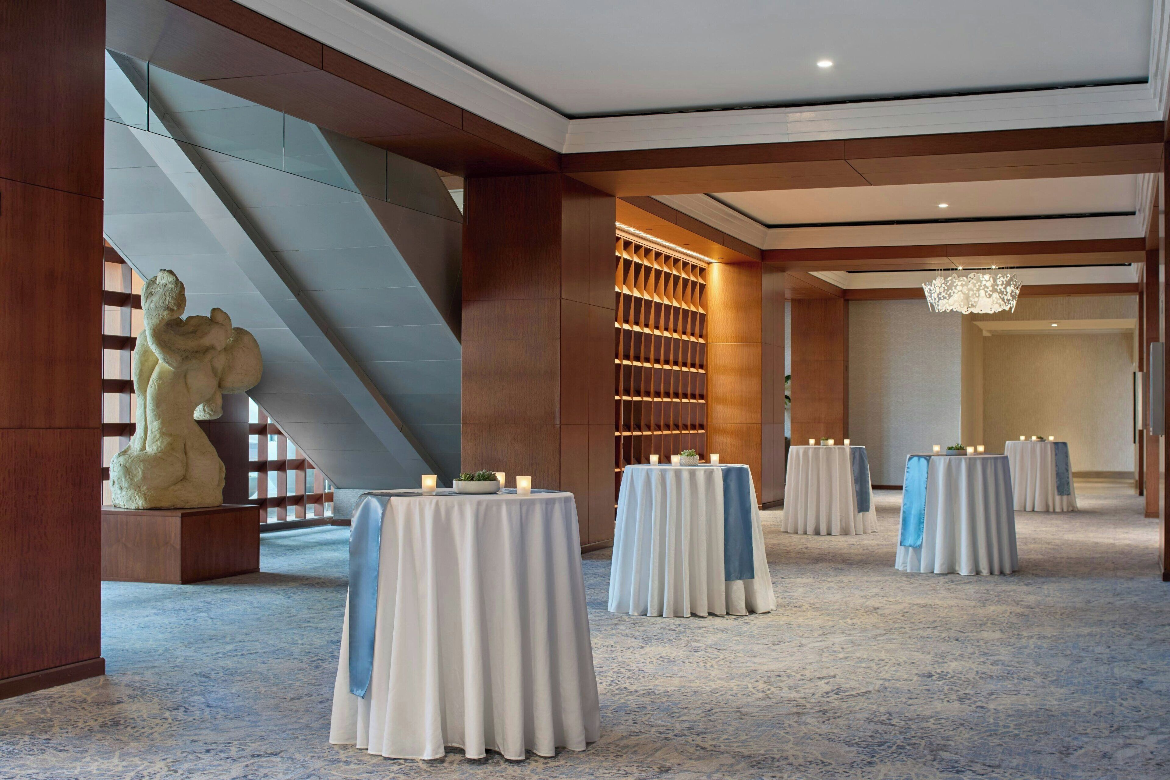 A spacious hotel lobby with a modern design, featuring a large staircase, a decorative sculpture, and multiple round tables set up for events.