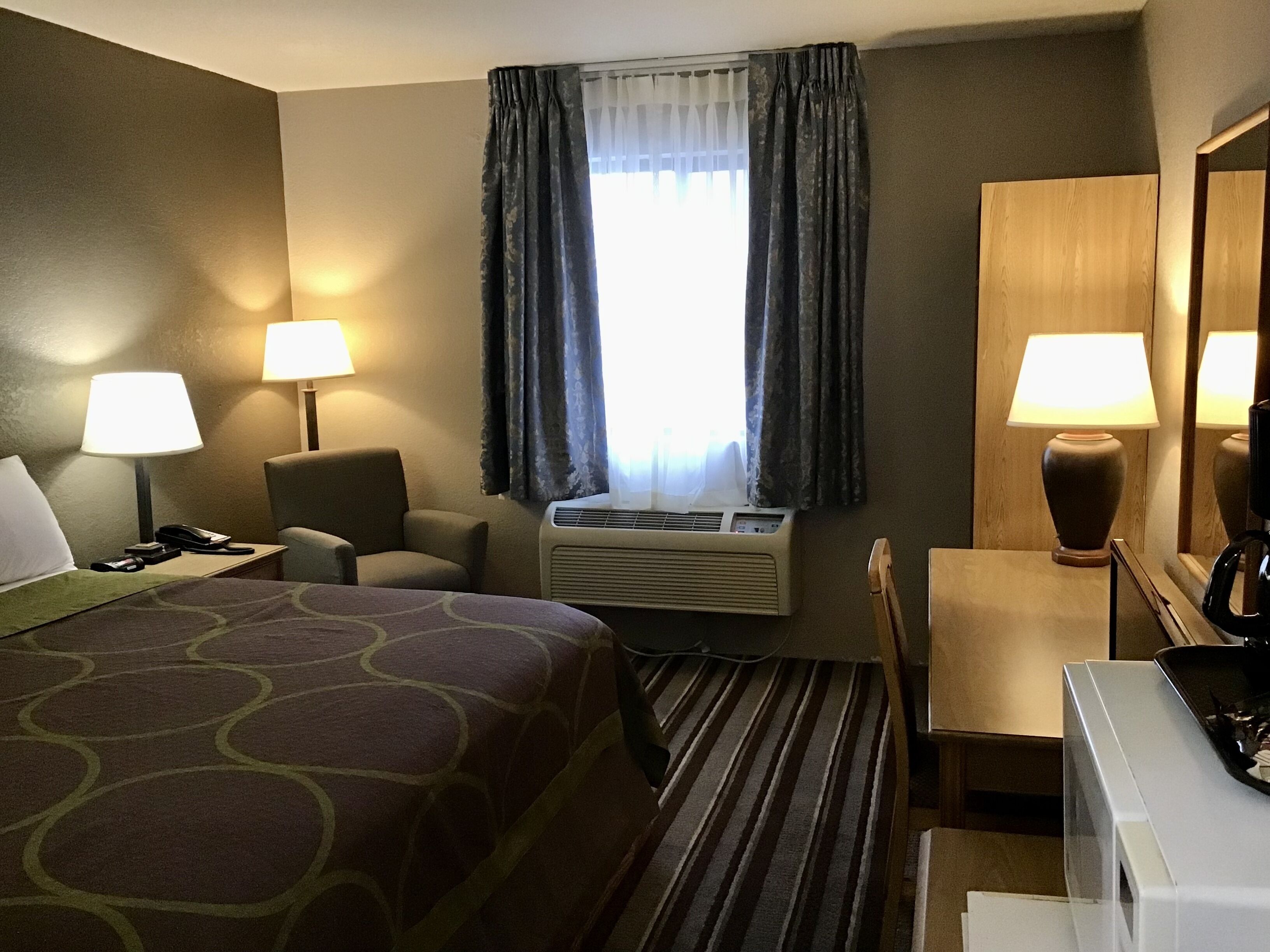 Et hotelværelse med en seng, to stole, et skrivebord, en lampe, aircondition og et vindue med gardiner.