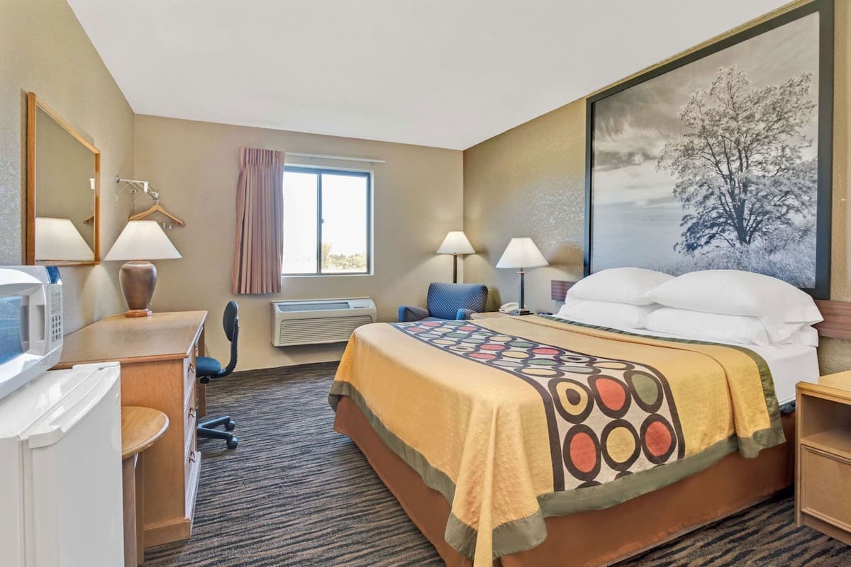 A hotel room with a bed, two bedside lamps, a window with curtains, a microwave, a desk with a telephone, and a large framed picture on the wall.