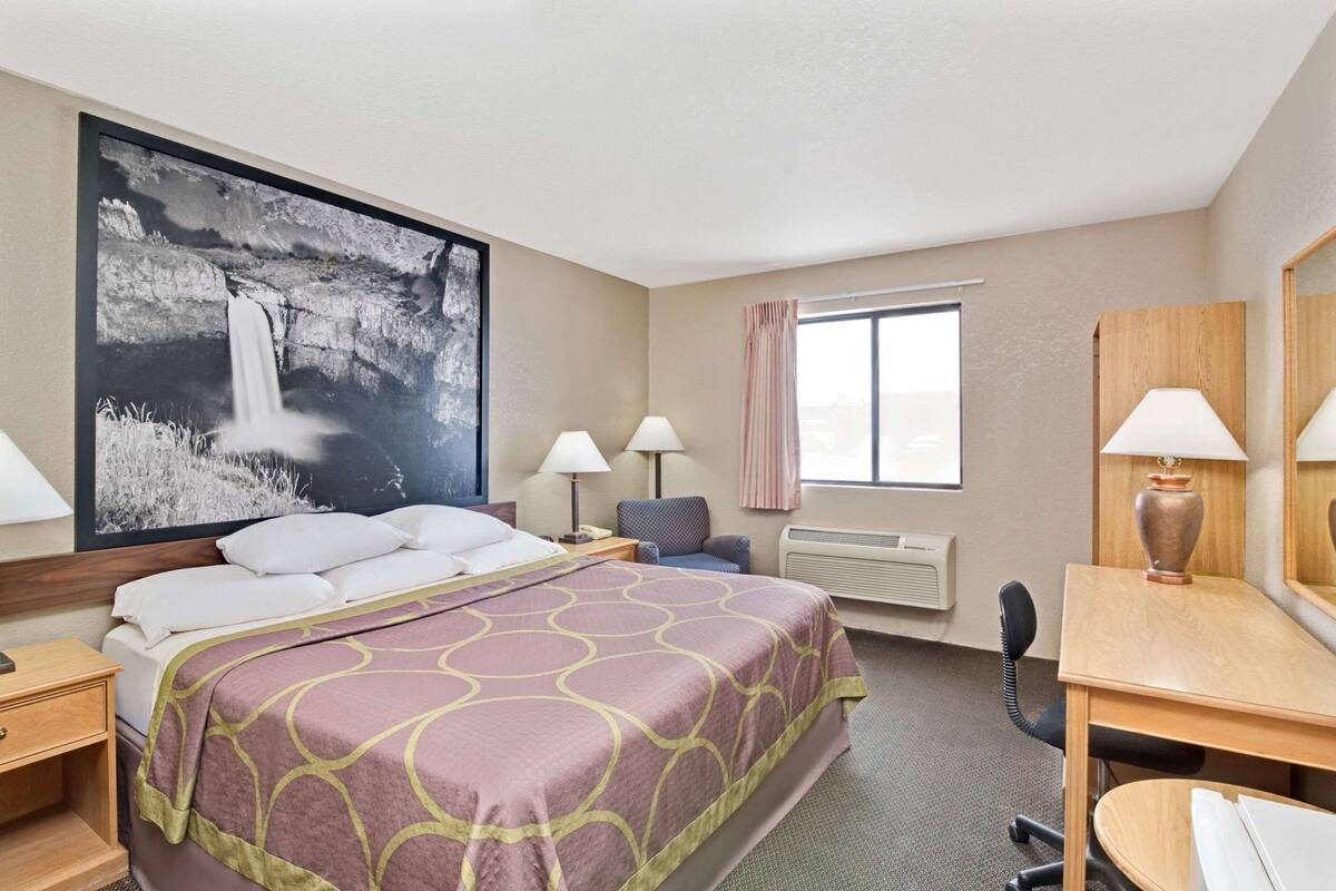 A hotel room with a large bed, two bedside tables with lamps, a desk with a chair, a large window, and a wall-mounted air conditioner.