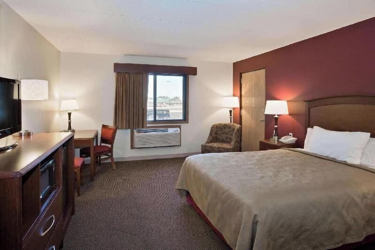 A hotel room with a bed, a desk with a chair, a television, a window with curtains, and an air conditioning unit.