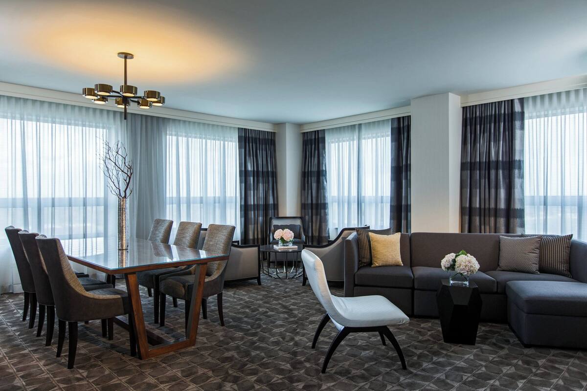 A modern living room with a dining area, featuring a glass-top table, dark sofas, and a patterned carpet.