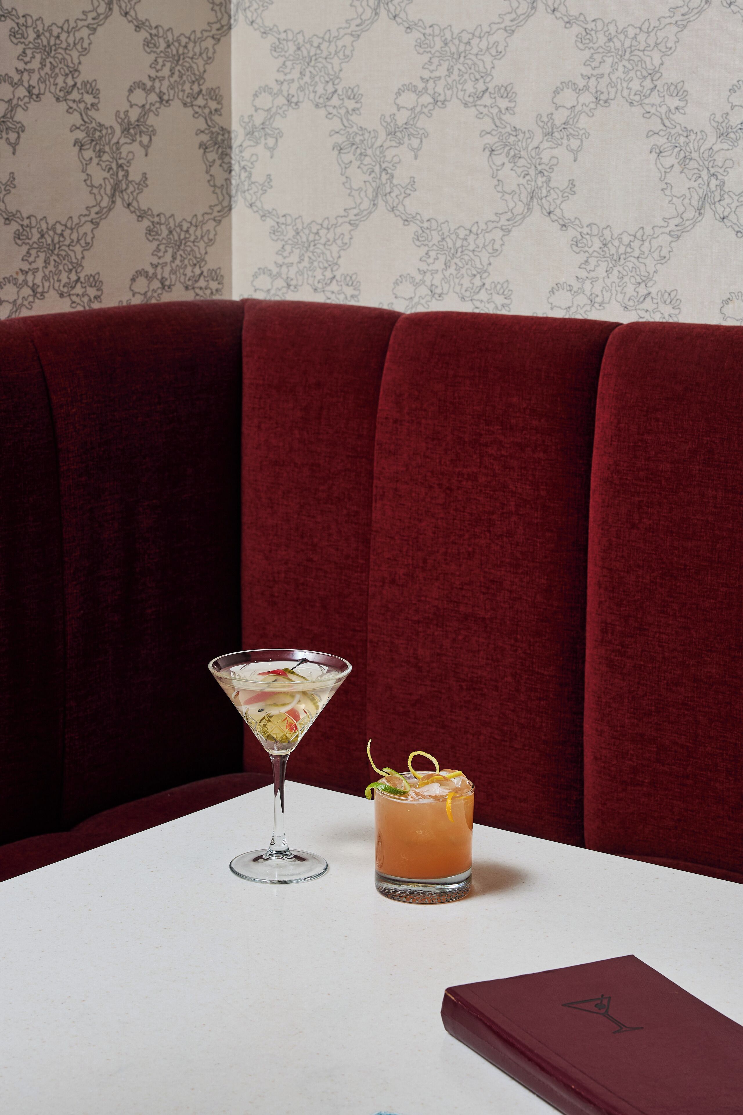 A table with a martini glass and a cocktail with a lemon twist, set against a red upholstered booth and a patterned wallpaper background.