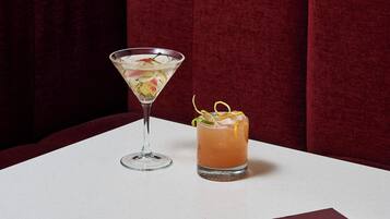 A table with a martini glass and a cocktail with a lemon twist, set against a red upholstered booth and a patterned wallpaper background.