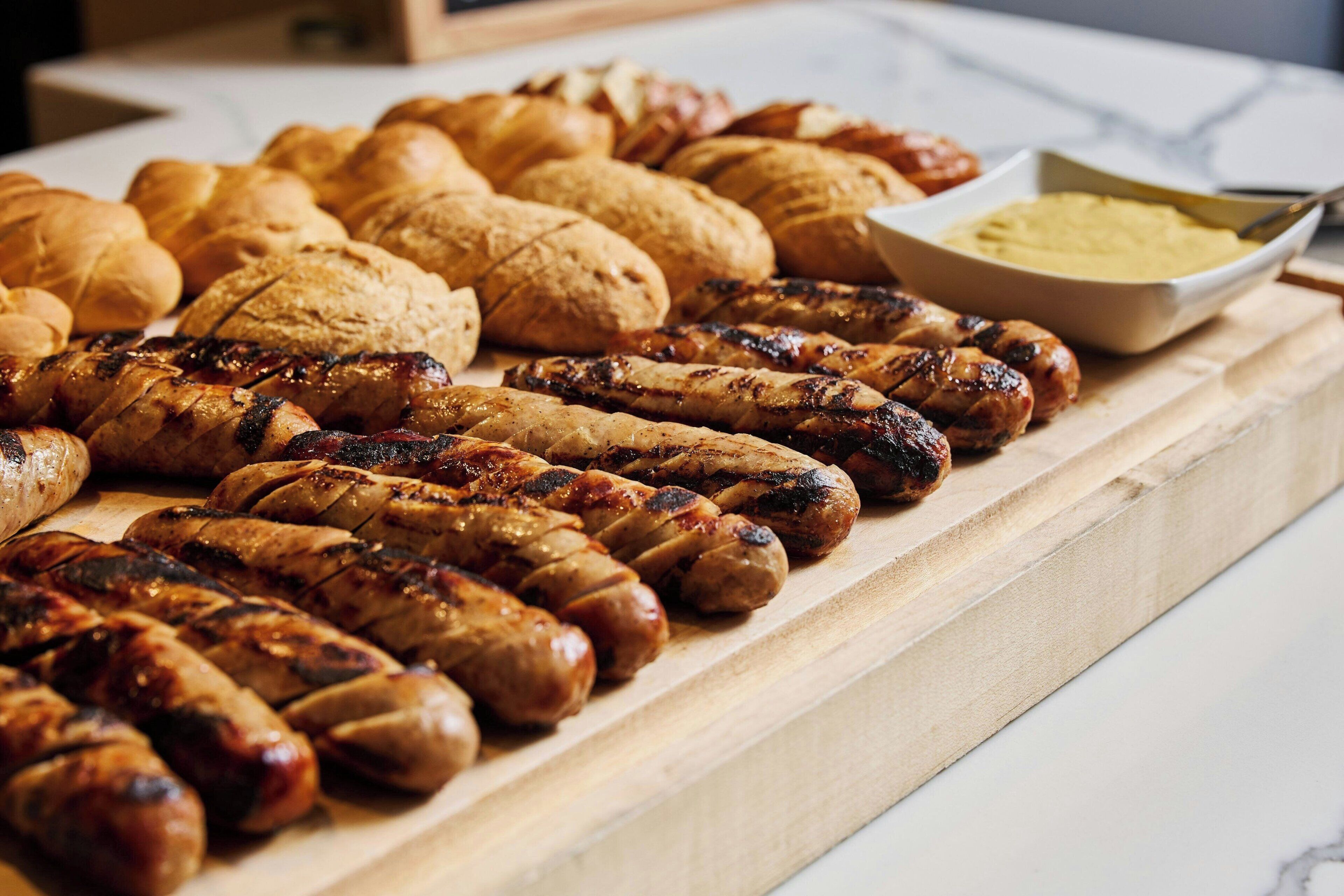 A wooden board with grilled sausages, croissants, and a bowl of mustard.