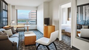 Habitación de hotel con sofá, escritorio, silla, cama, televisor y un amplio ventanal con vistas a la ciudad.